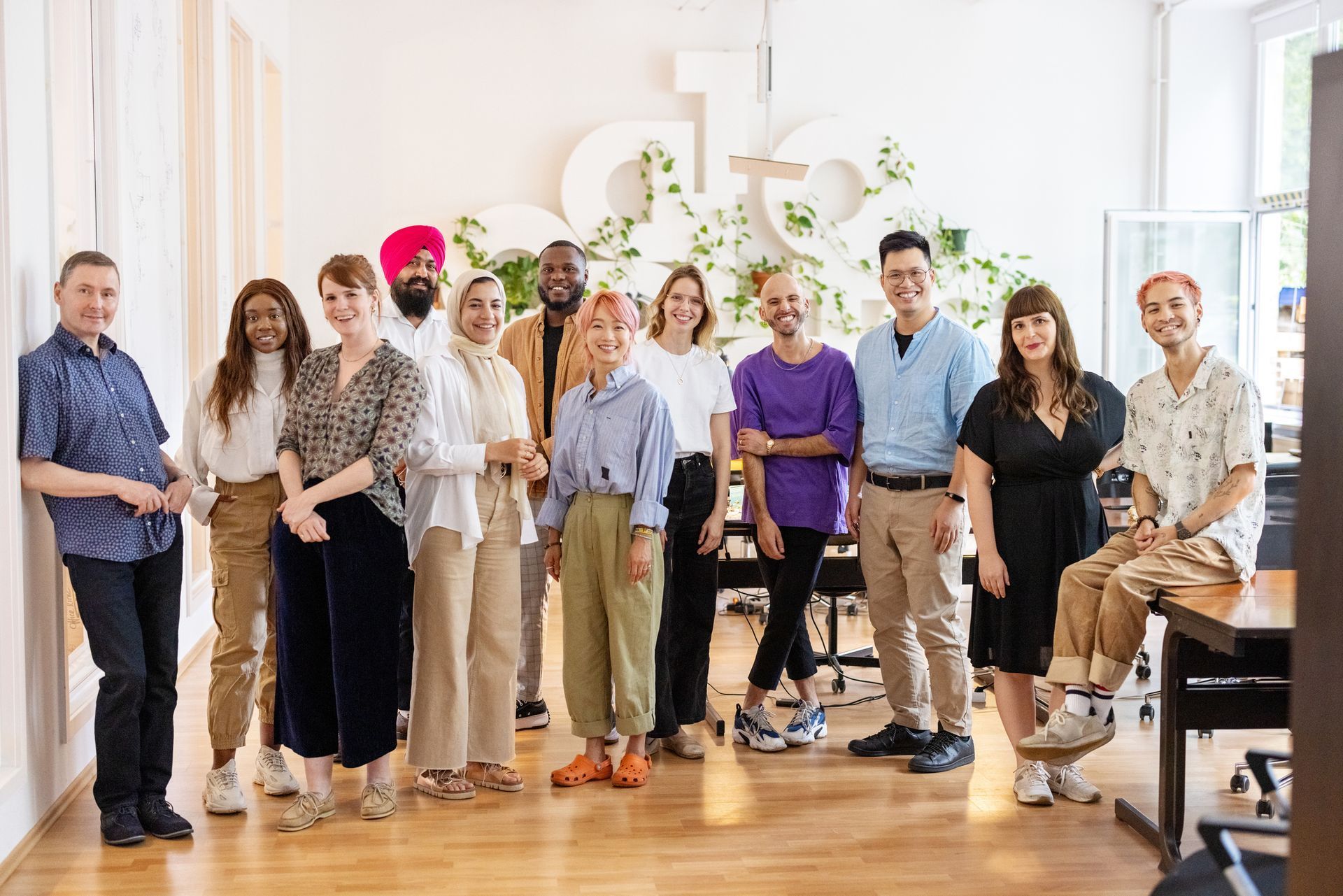 Group of diverse people in a brightly lit office, smiling and posing. Green plant decor on the wall.