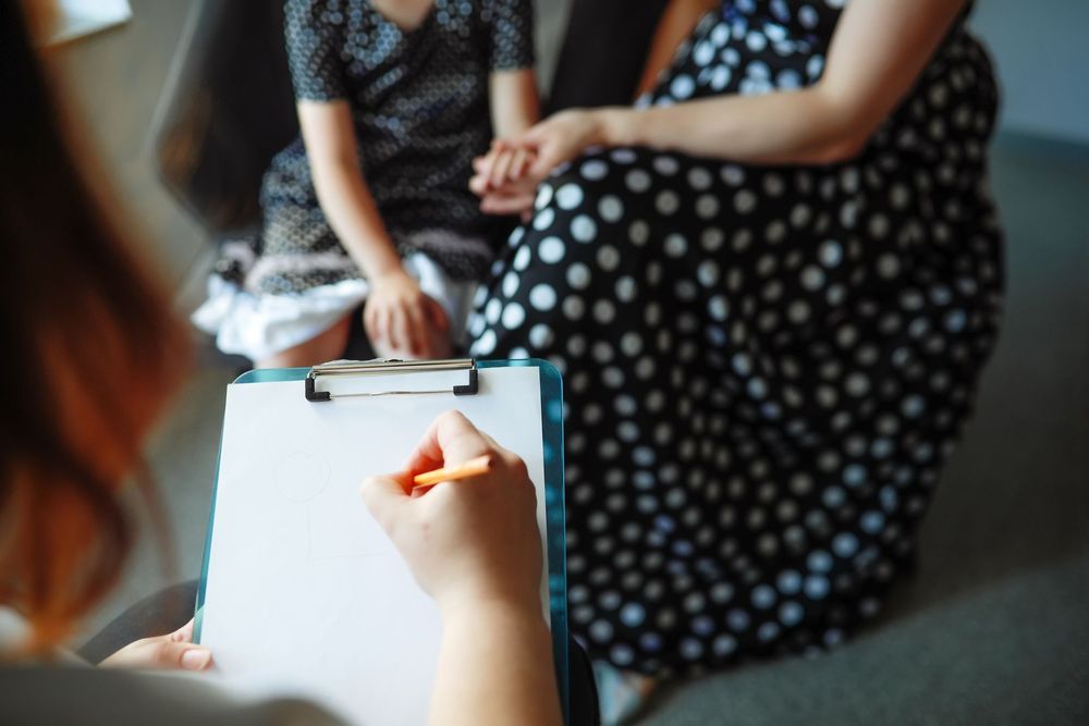 A person writing on a clipboard, meeting with a child and an adult in a consultation setting.