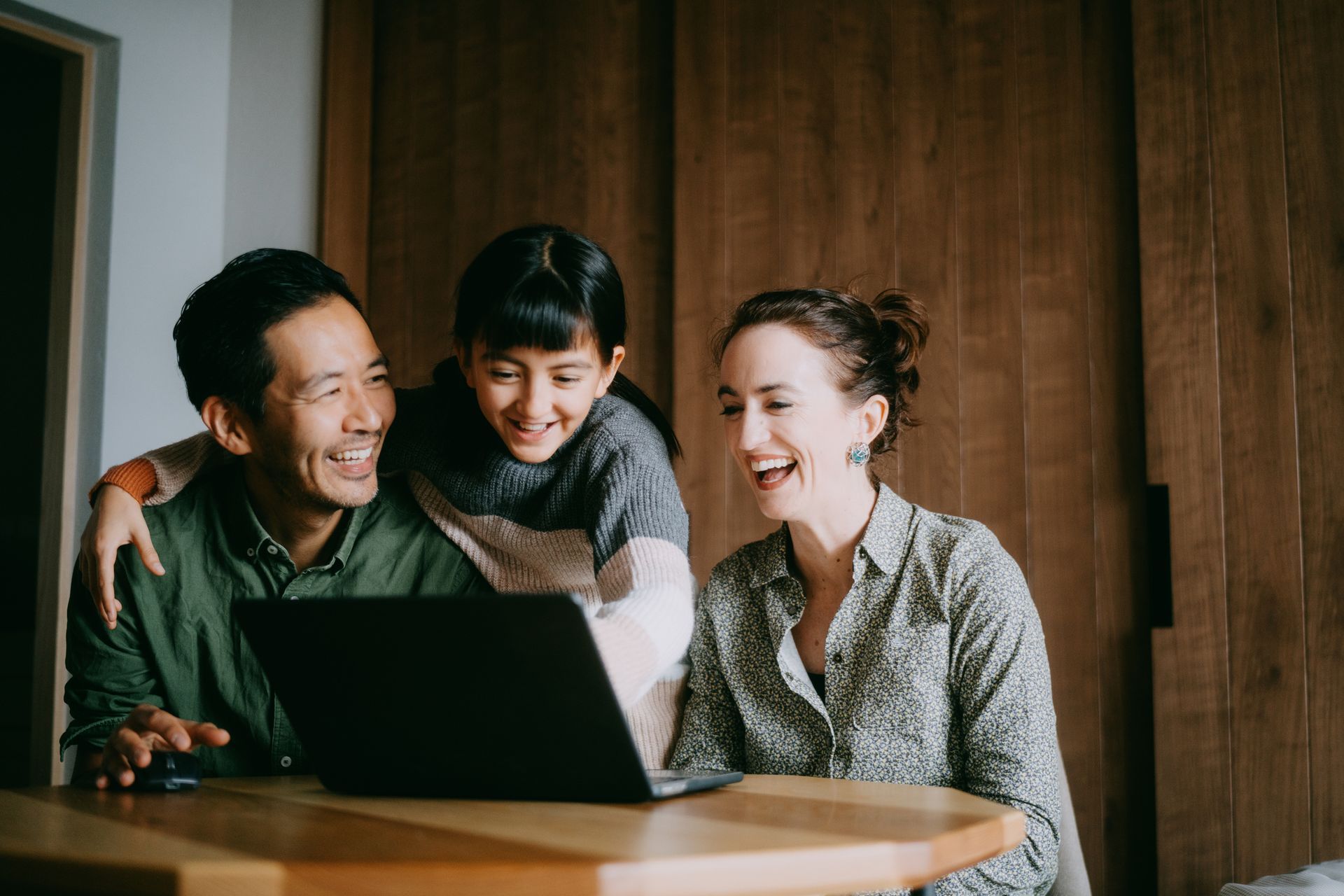Family of three smiling, gathered around a laptop on a wooden table.