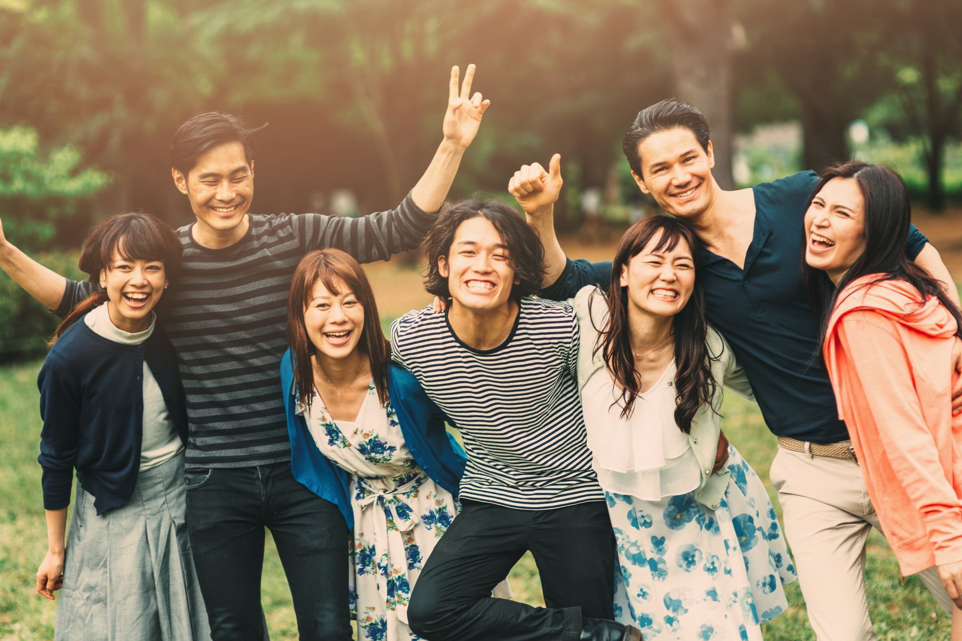 Group of smiling people in a park, arms around each other. Sunlit scene.