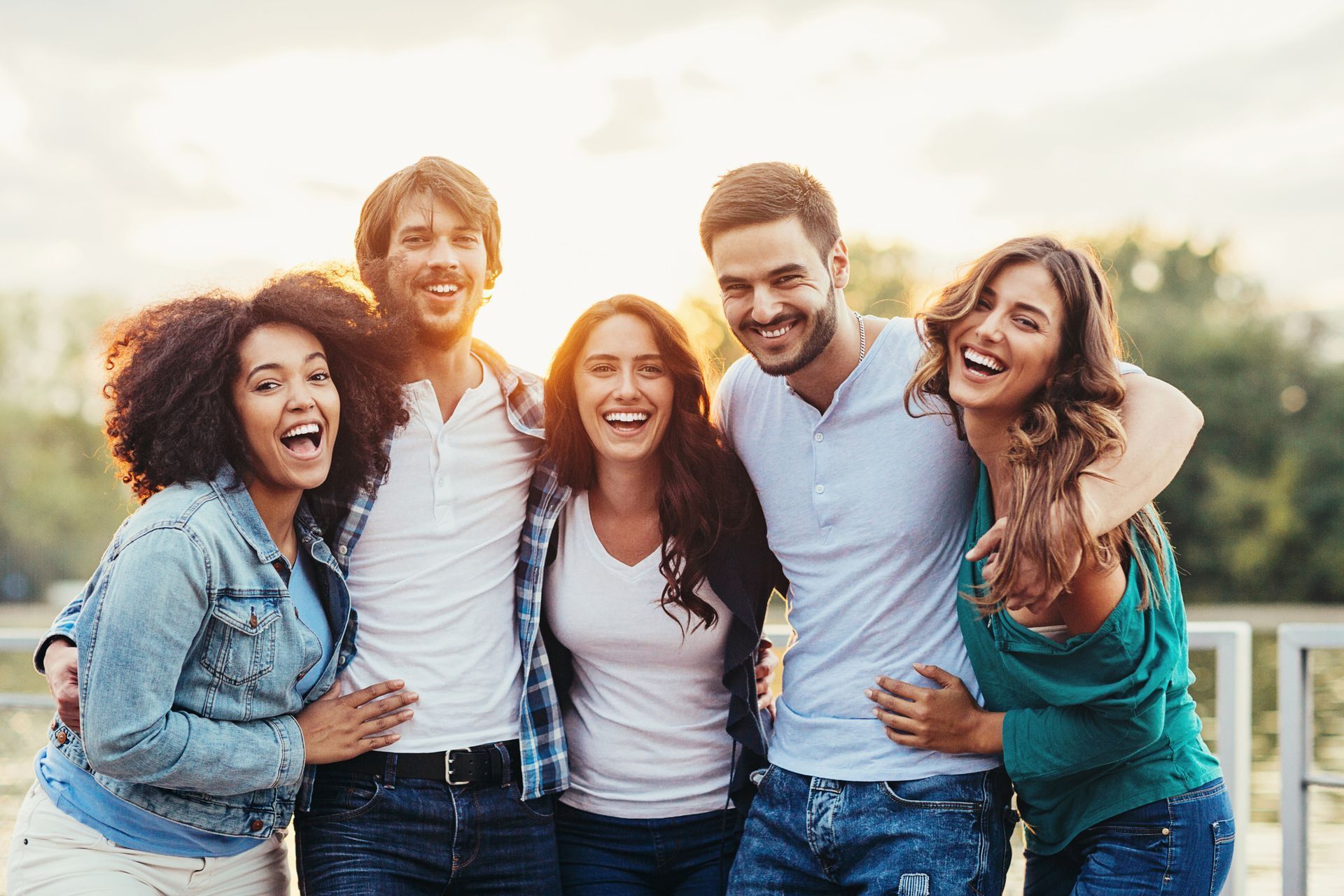 Five smiling people with arms around each other, laughing outdoors in sunlight.