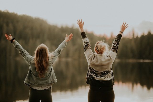 Two people with arms raised by a lake, trees in the background, golden light.