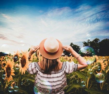 Woman in hat and striped shirt in sunflower field, looking up at the sky. Bubbles float in the air.