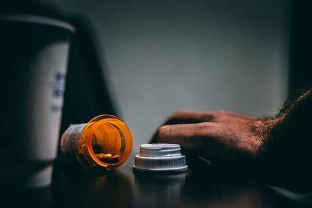 Open prescription bottle next to a hand and a cup, indoors.
