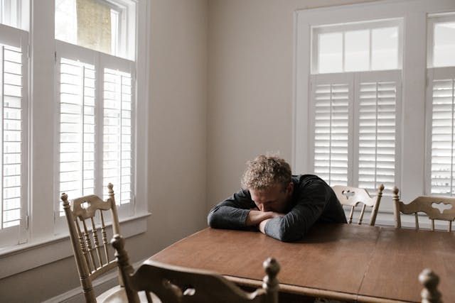 Man with head on hands, at a wooden table in a room with white shutters; looks distressed.