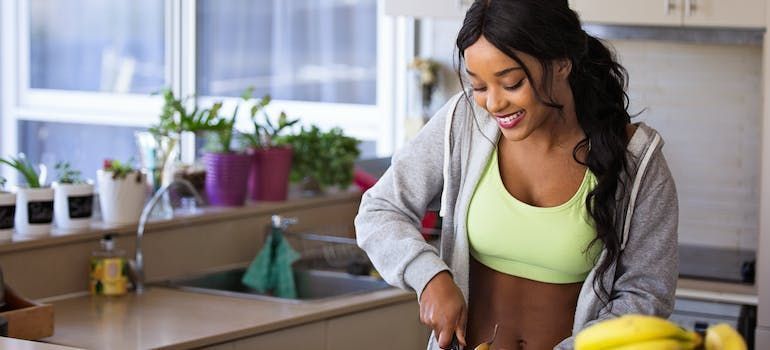 Woman in a kitchen slicing fruit; smiling, wearing a sports bra and jacket.