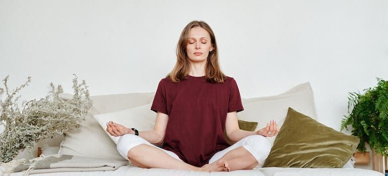 Woman meditates on a couch with closed eyes, wearing a red shirt and white pants.