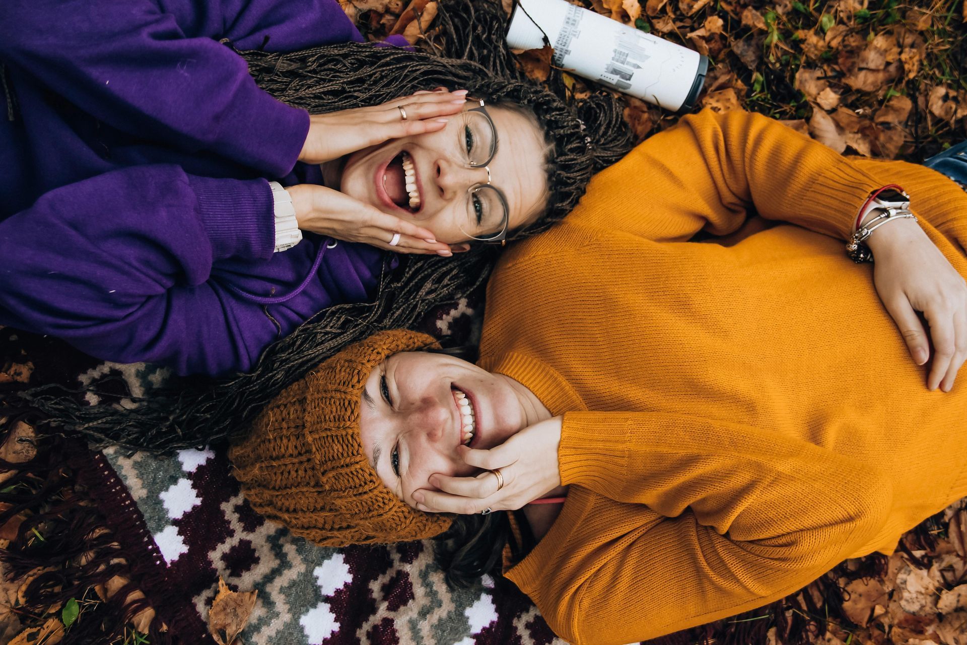 Two smiling people lying on a blanket, surrounded by leaves. One wears orange, the other purple.