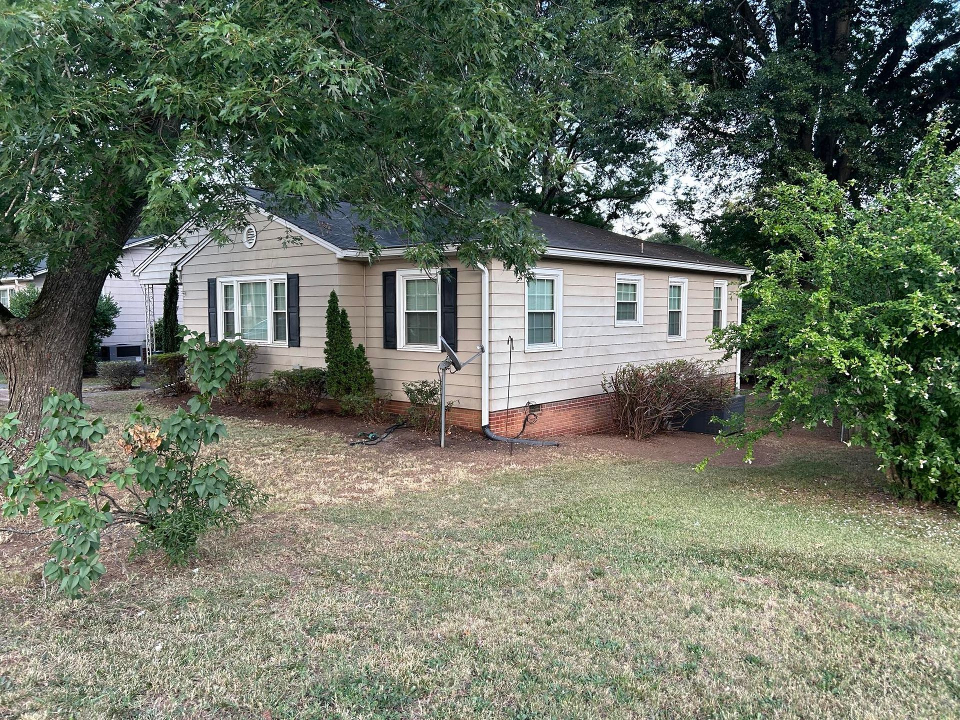 A one-story beige house with black shutters and a dark roof, surrounded by trees and grass.
