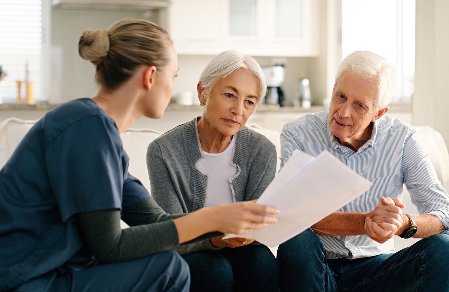 An elderly couple is sitting on a couch talking to a nurse while looking at a piece of paper.