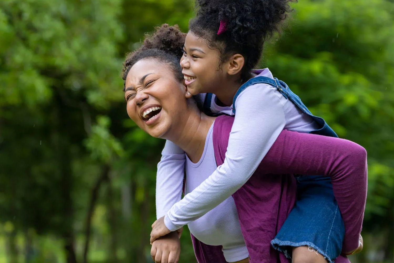 A woman is giving a little girl a piggyback ride in a park.