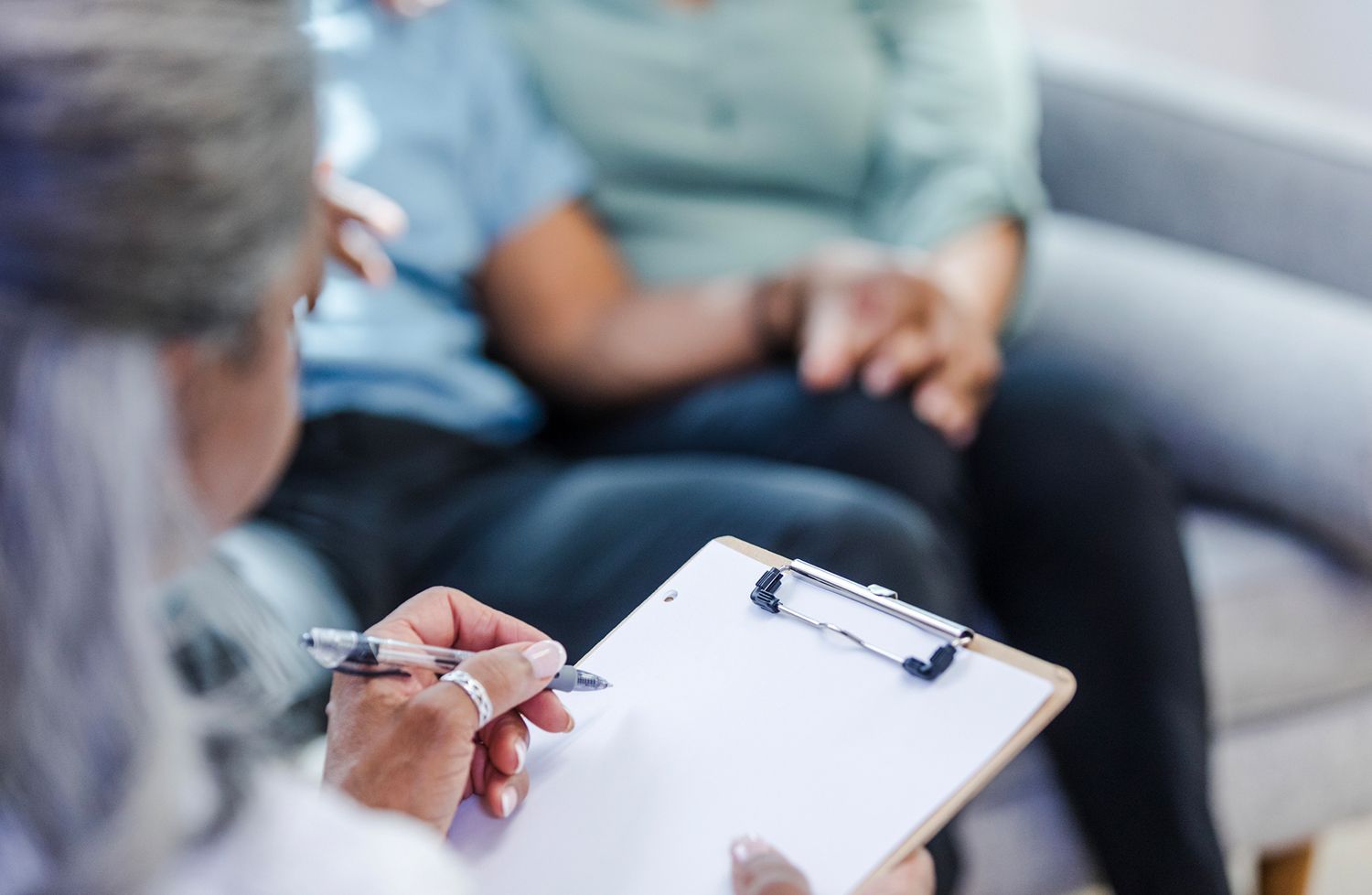 A woman is writing on a clipboard while a man and woman sit on a couch.