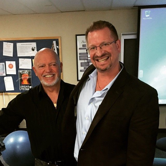 Two men smiling, one bald in black shirt, the other in glasses and blazer, indoors with a bulletin board and screen.