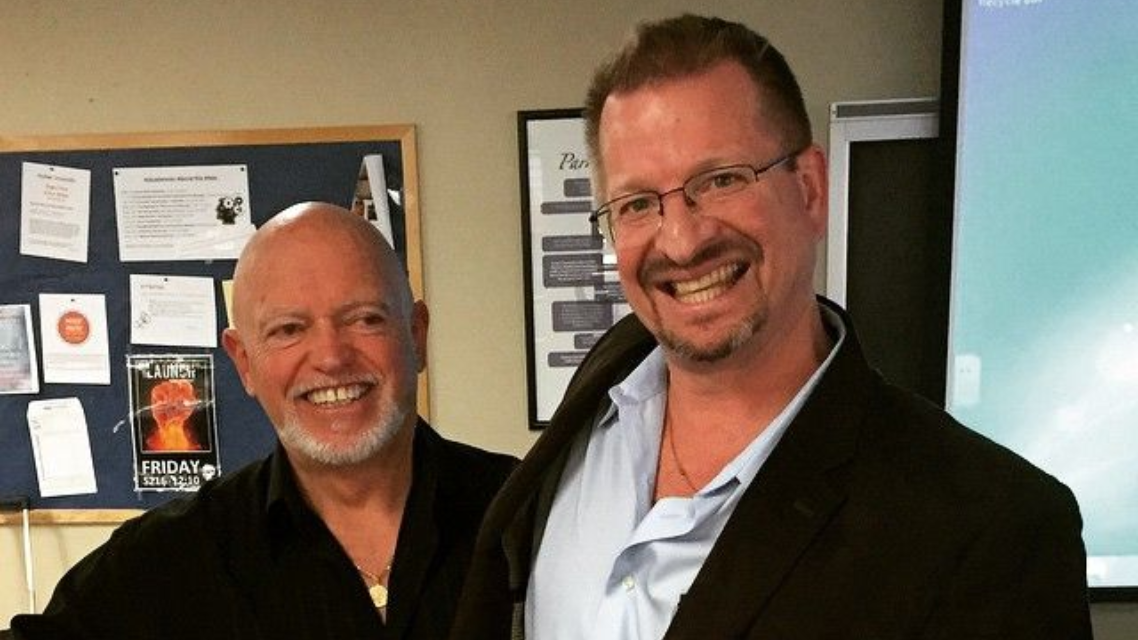 Two men smiling, one bald in black shirt, the other in glasses and blazer, indoors with a bulletin board and screen.