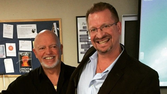 Two men smiling, one bald in black shirt, the other in glasses and blazer, indoors with a bulletin board and screen.