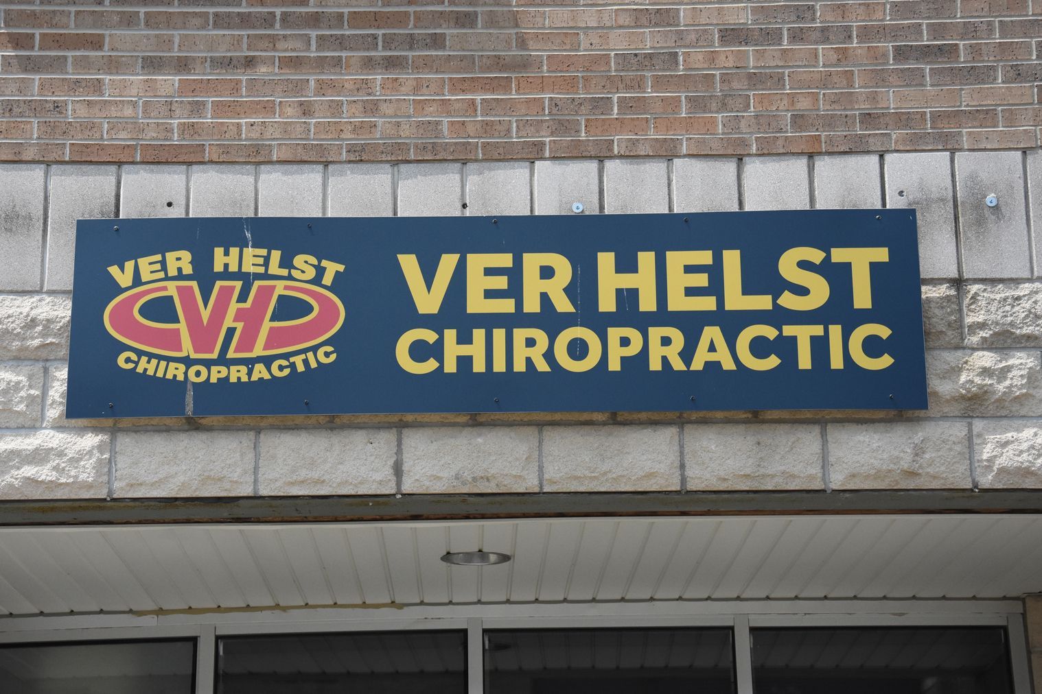 Sign for Ver Helst Chiropractic, blue background, white lettering, red logo, above a building entrance.