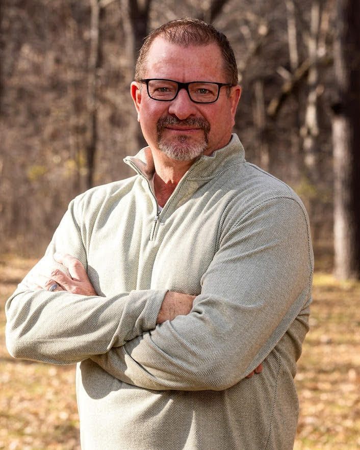 Man with glasses and folded arms, wearing a green quarter-zip in an outdoor setting.