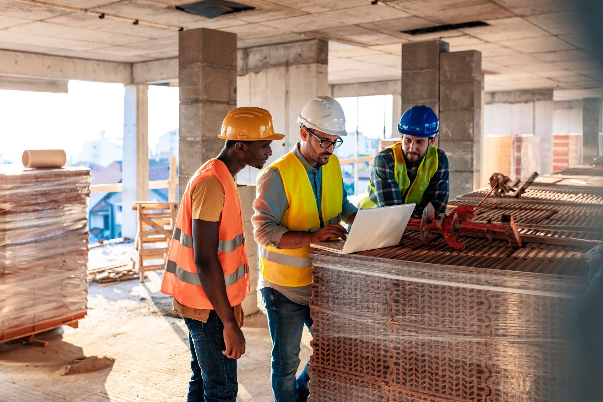 Construction workers looking at a laptop on a job site, discussing plans, wearing hard hats and safety vests.