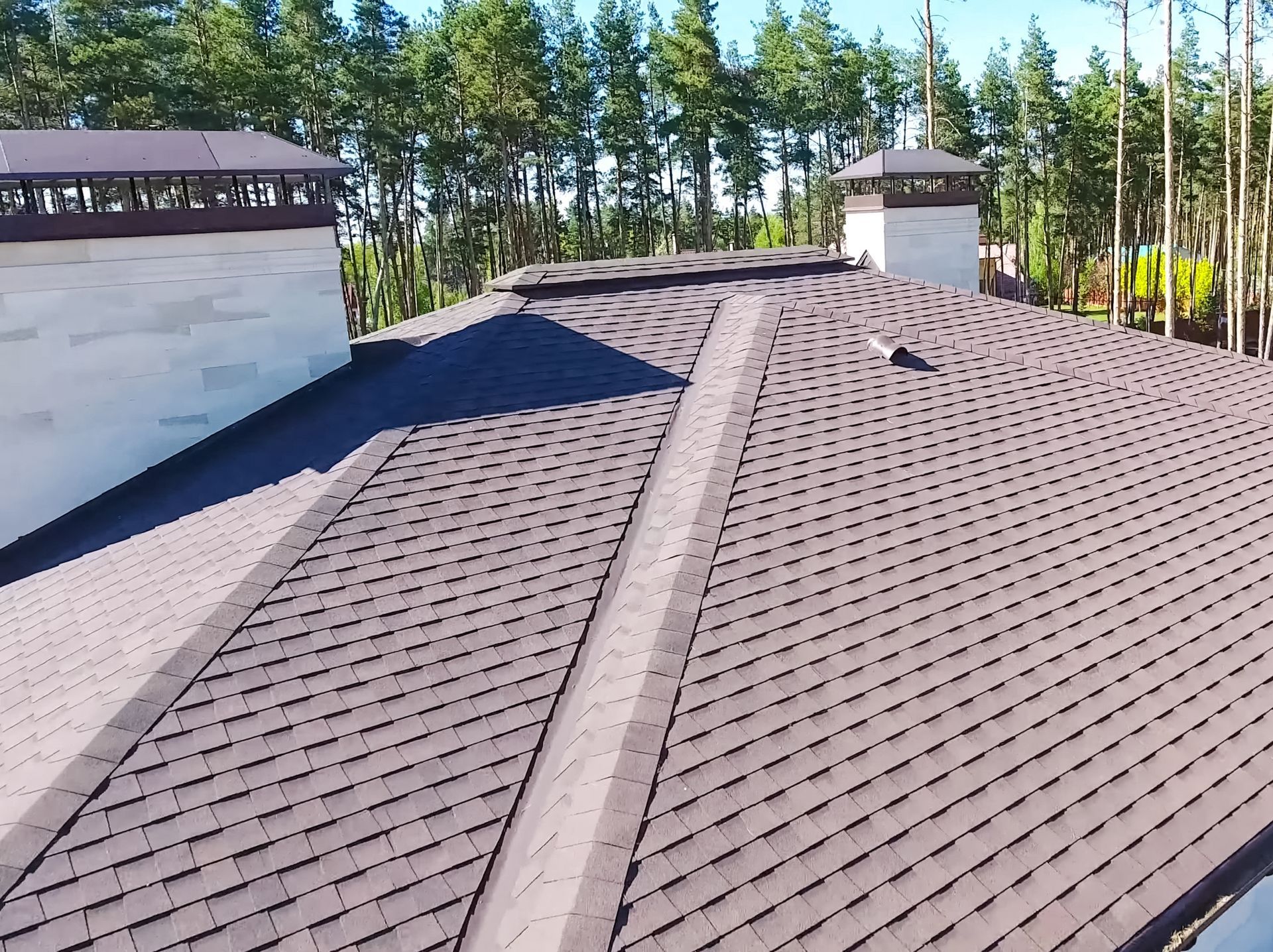 Brown shingled roof with chimneys against a backdrop of trees and a clear sky.