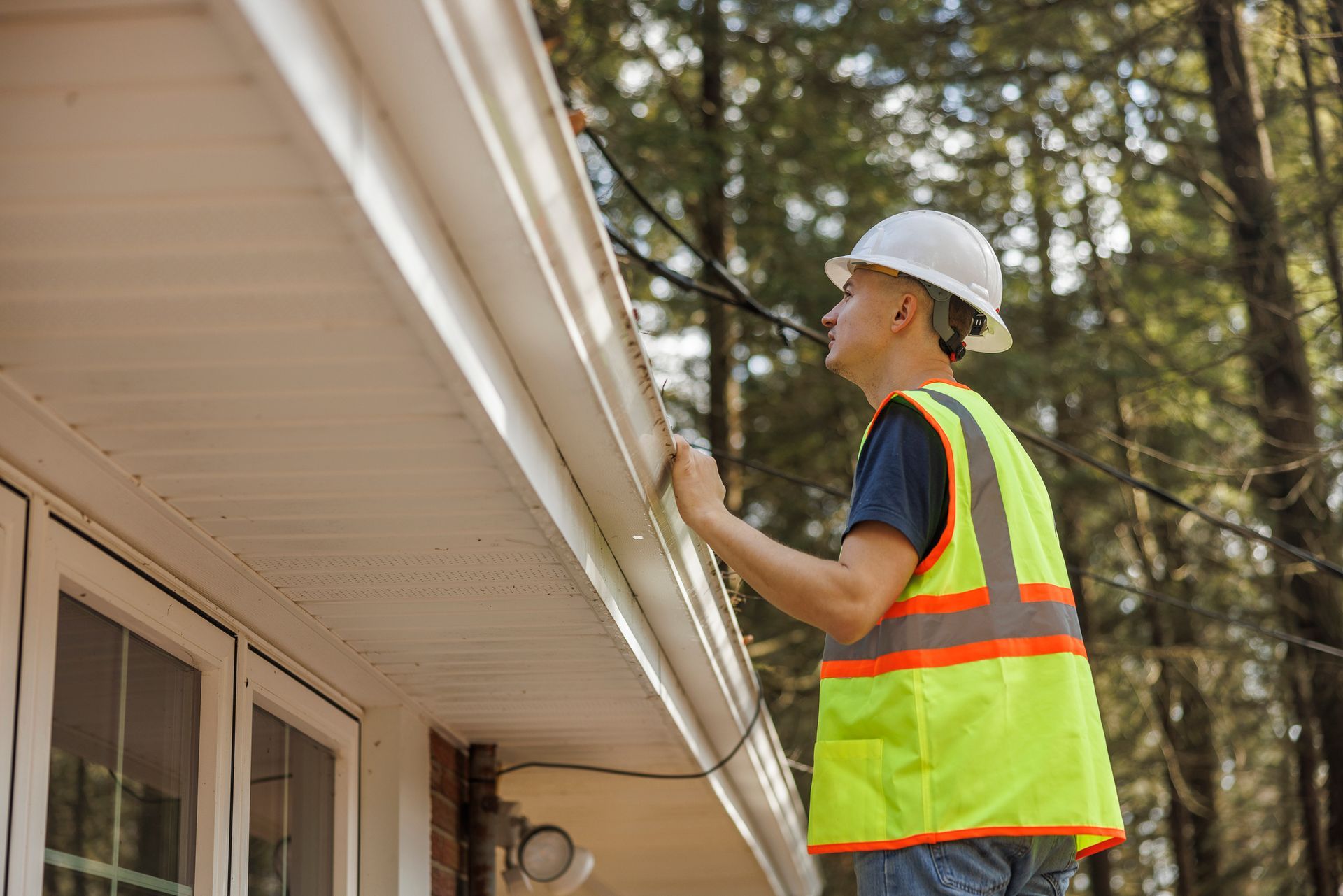 Person in hard hat and safety vest inspecting a house gutter.