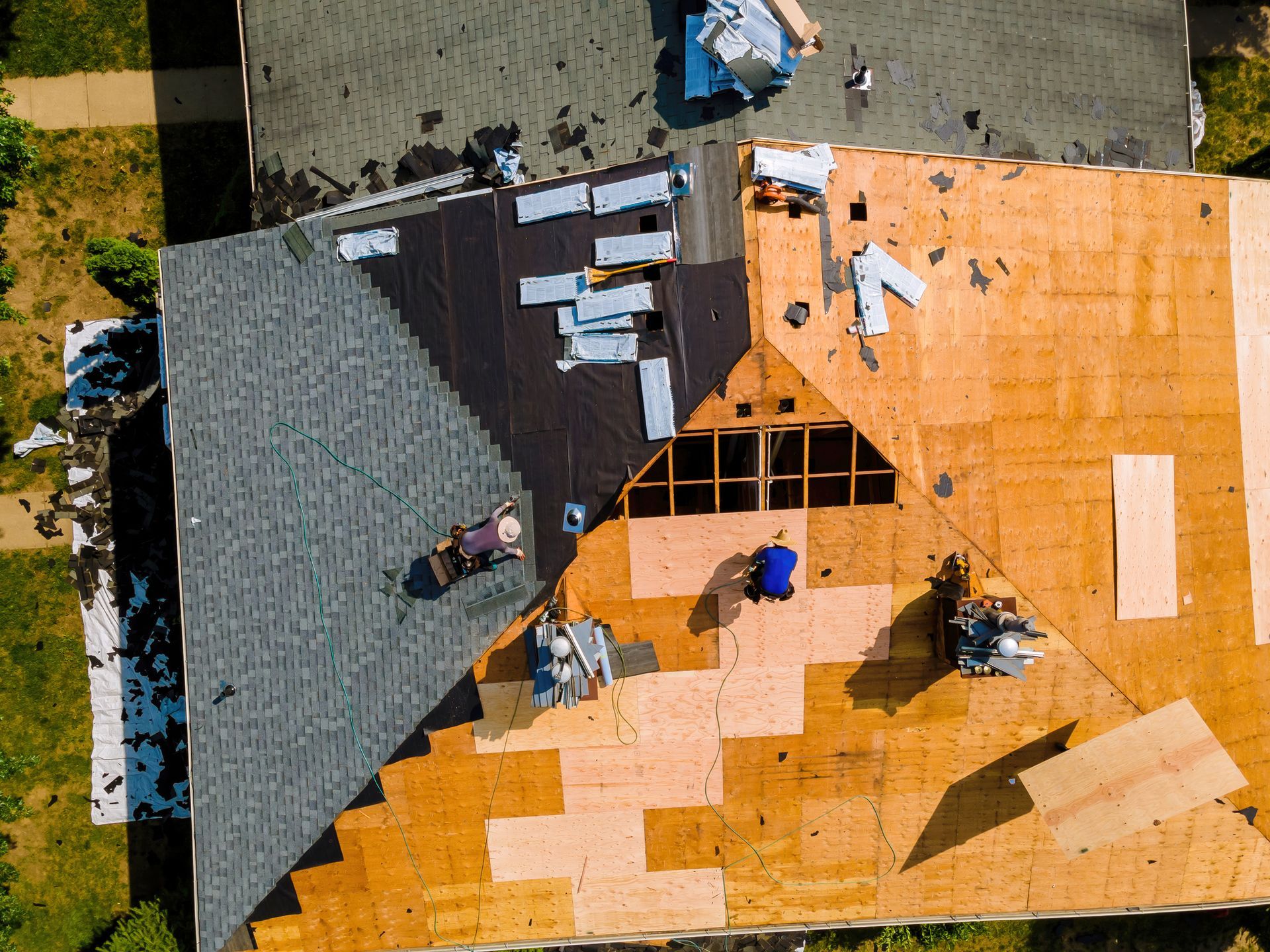 Roofers working on a house roof. Various stages of construction visible: removing old shingles, installing new materials.