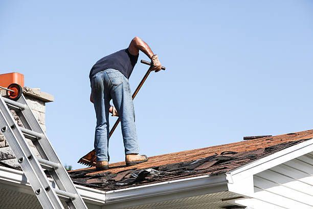 Man on roof removing shingles with a shovel, ladder and chimney visible.