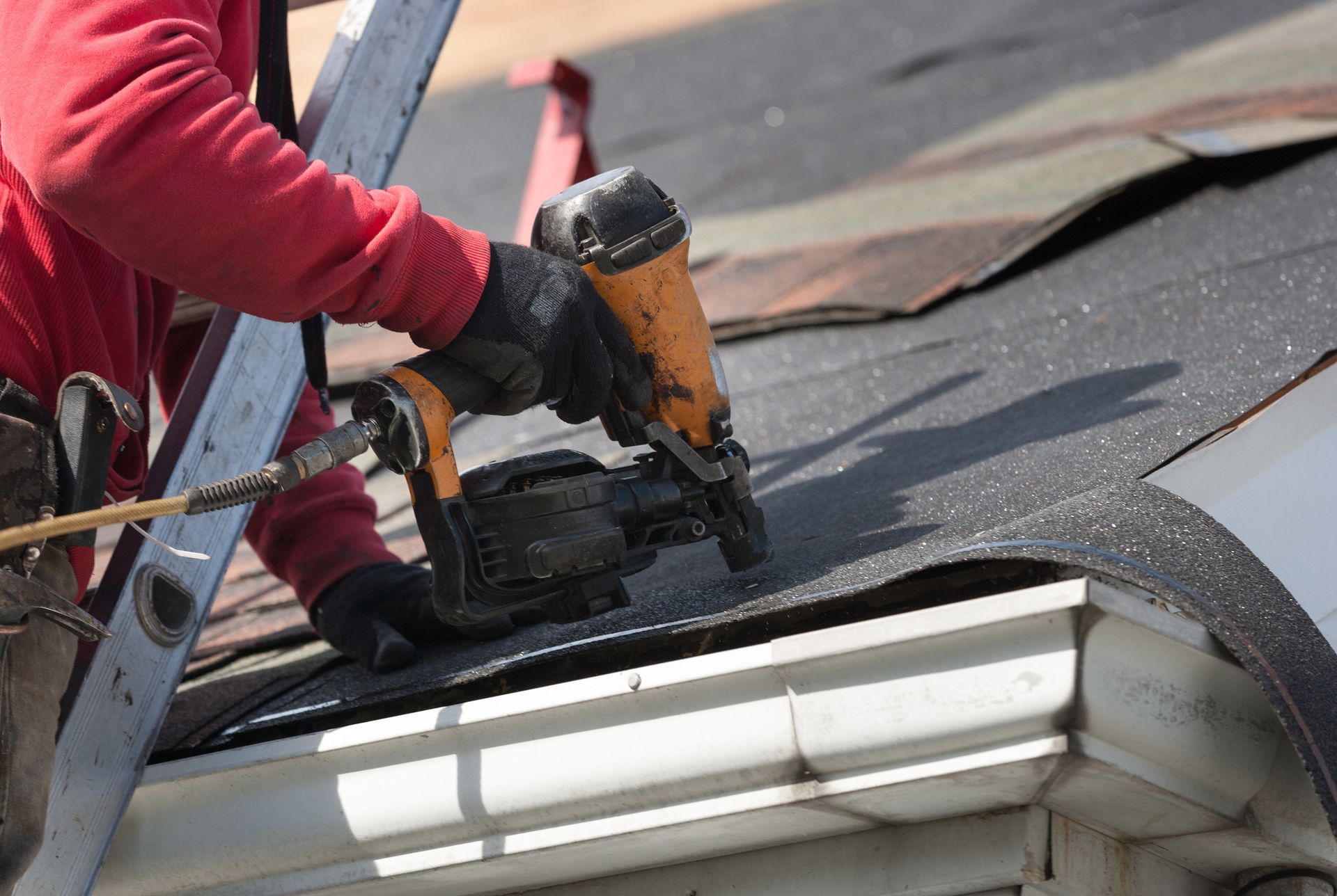 Roofer in red shirt using a nail gun on a roof, near a gutter, with ladder in background.