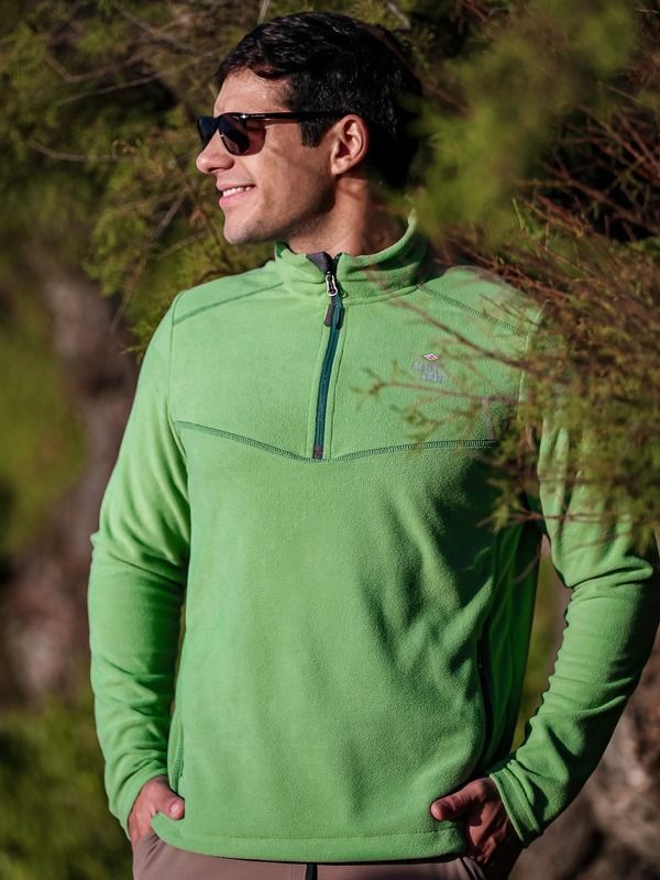 Un hombre que lleva gafas de sol y un suéter verde está parado frente a un árbol.