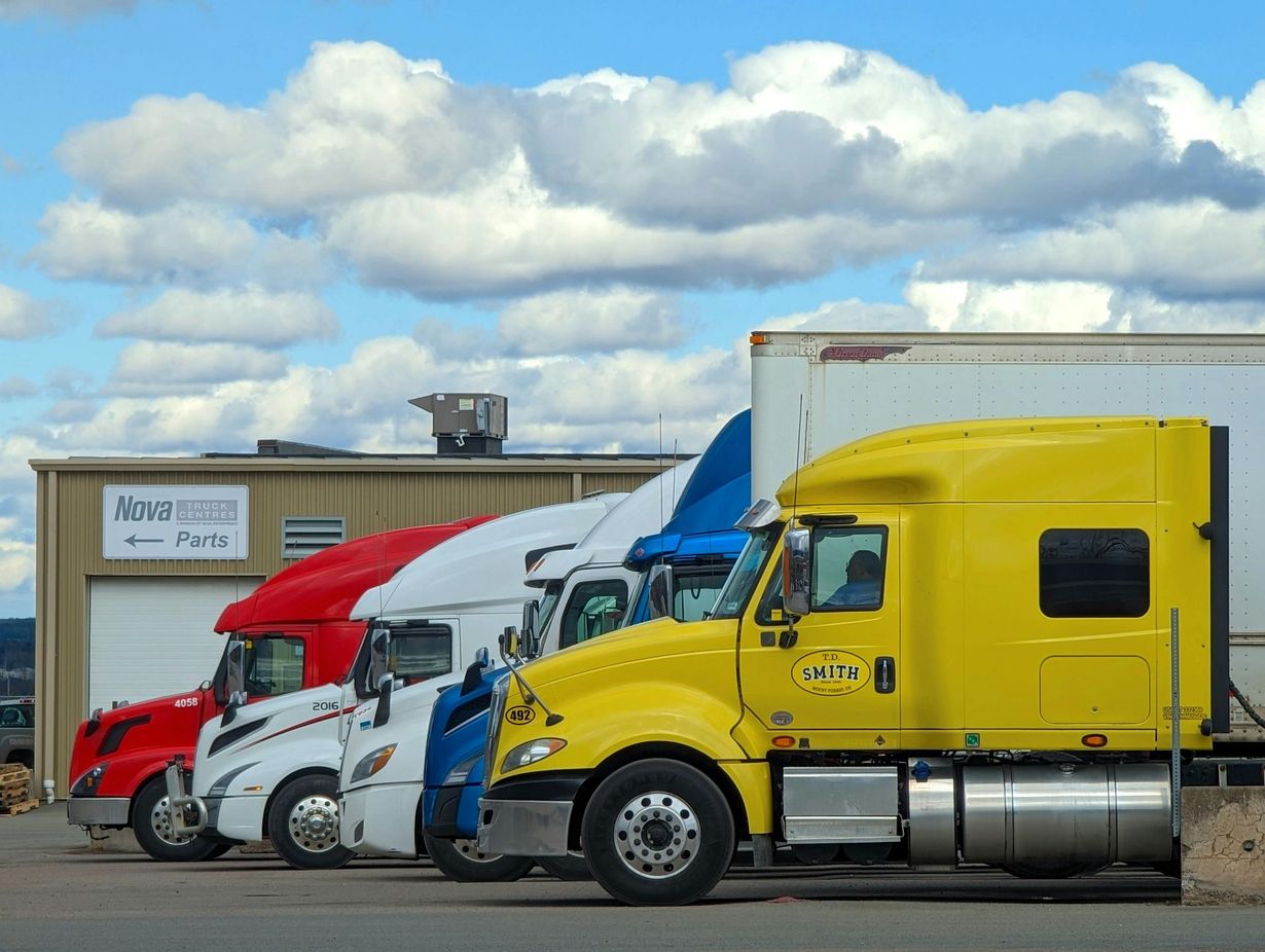 A line of semi-trucks in yellow, blue, white, and red parked in front of a warehouse under a cloudy sky.