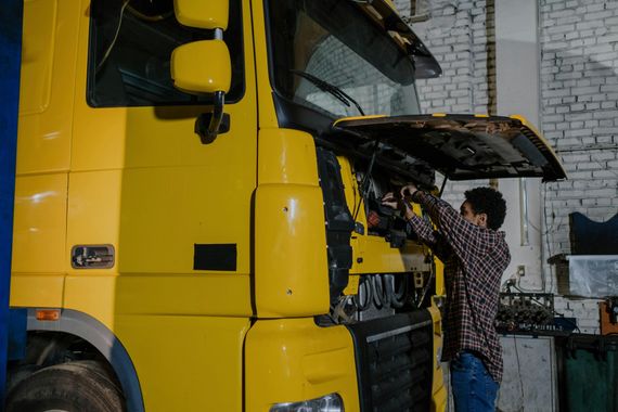 A person in a plaid shirt repairs the open engine compartment of a large yellow semi-truck inside a garage.