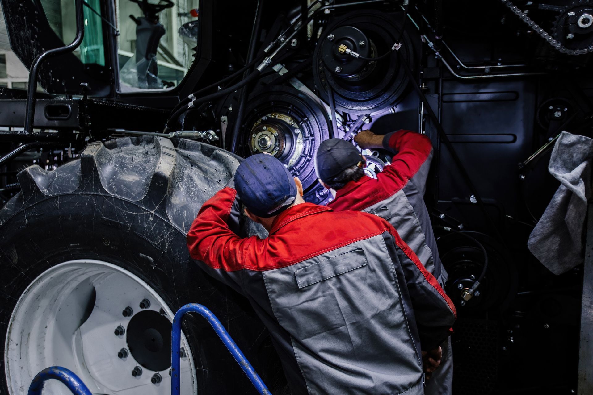Two workers in red and gray uniforms perform maintenance on the complex machinery of a large tractor indoors.