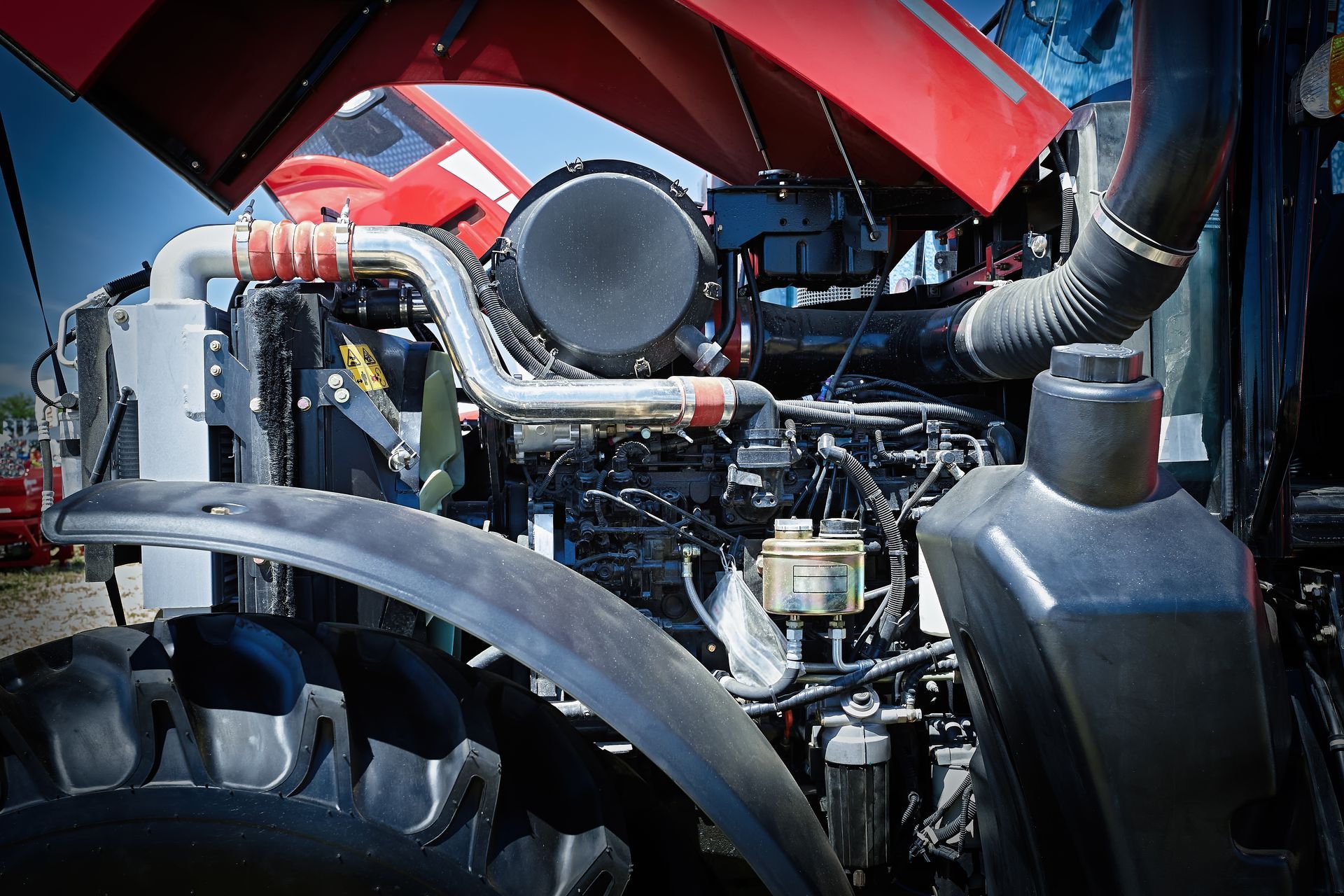 The open hood of a red tractor revealing its dark, complex engine, internal components, and parts of the front tire.