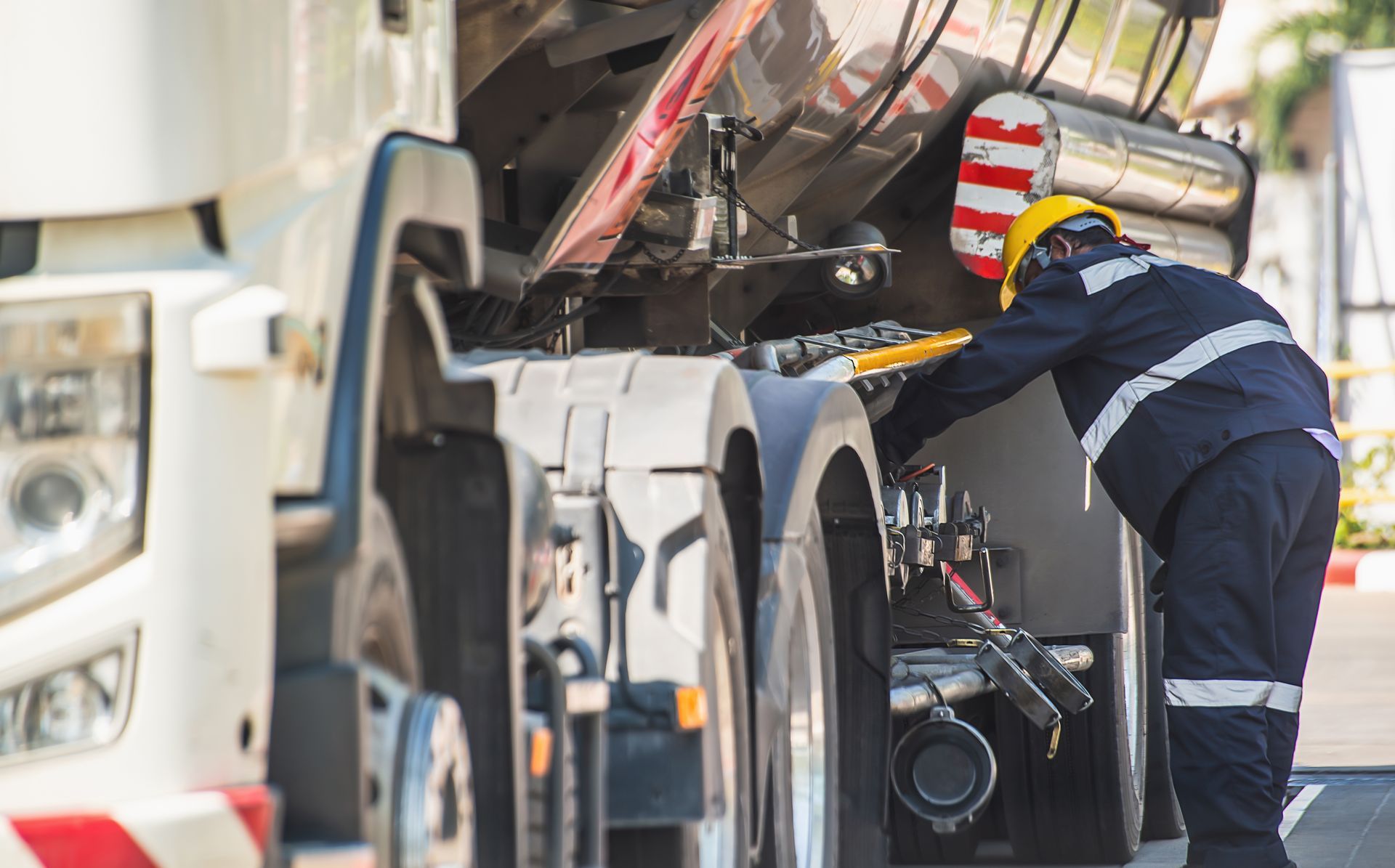 A worker in a high-visibility uniform and yellow hard hat inspects the piping underneath a fuel tanker truck.