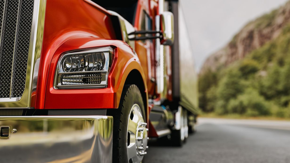 Close-up, low-angle view of a shiny red semi-truck parked on the side of a road with a blurred mountain background.