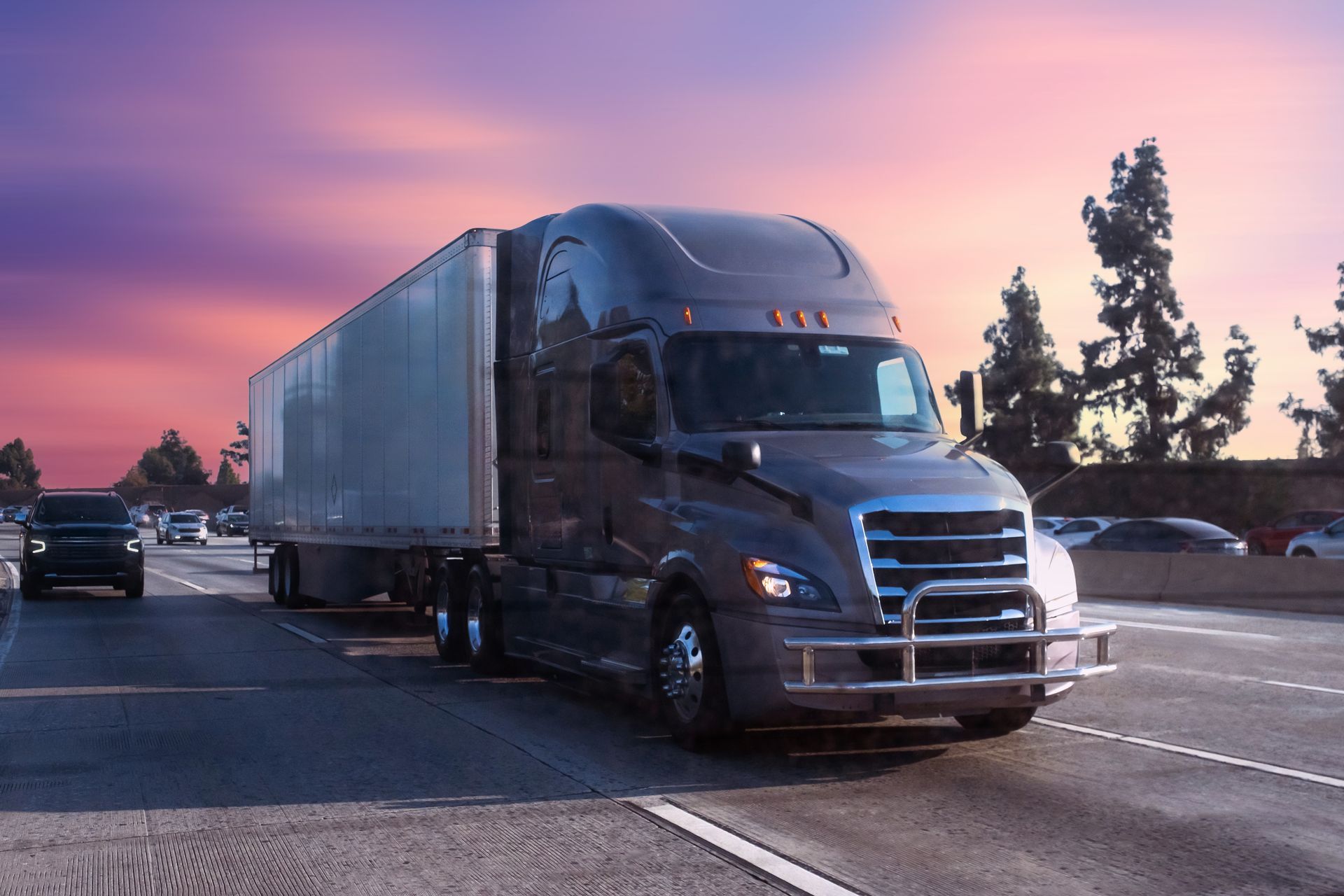 A semi-truck with a large trailer driving on a highway during a vibrant purple and orange sunset.