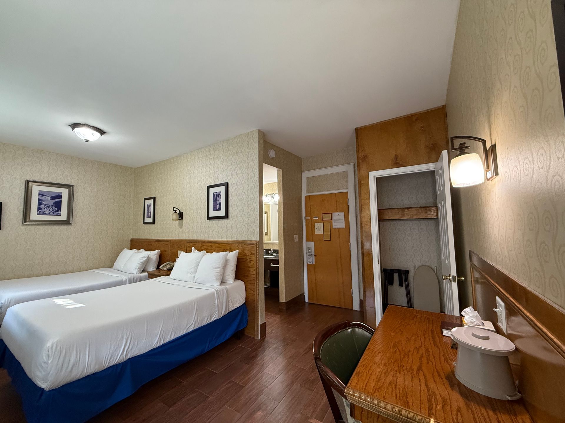 Hotel room with two twin beds, desk, and bathroom doorway. Beige walls, wood accents, and artwork.