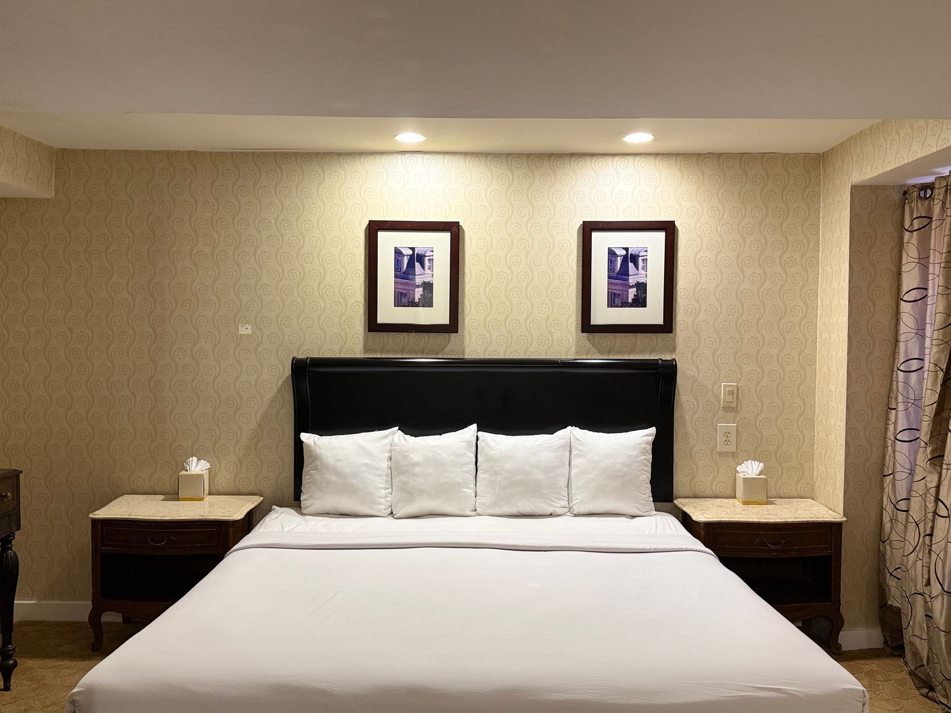 Hotel bedroom with a bed, nightstands, artwork, and patterned wallpaper.