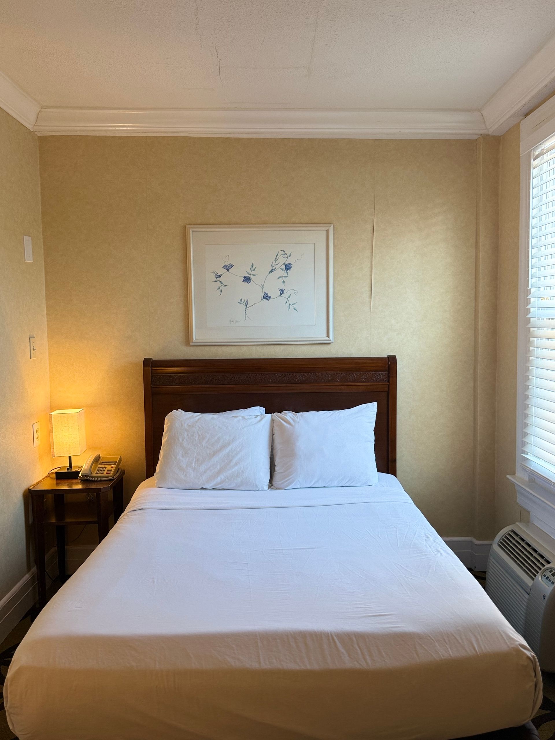 Bedroom with a made bed, nightstand, lamp, and framed art on the wall. Window on the right.
