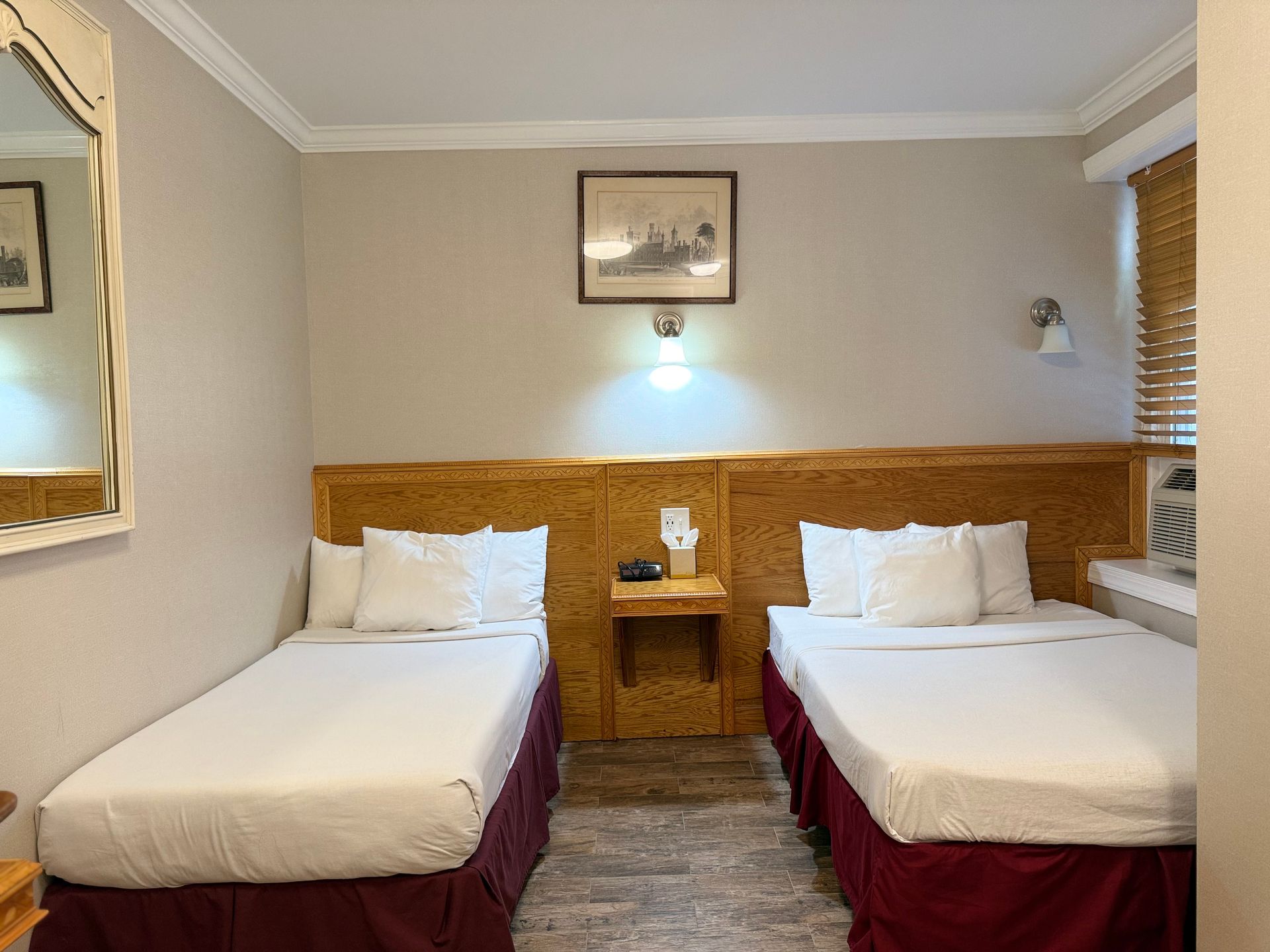Two twin beds with white sheets and burgundy skirts in a hotel room with tan walls.