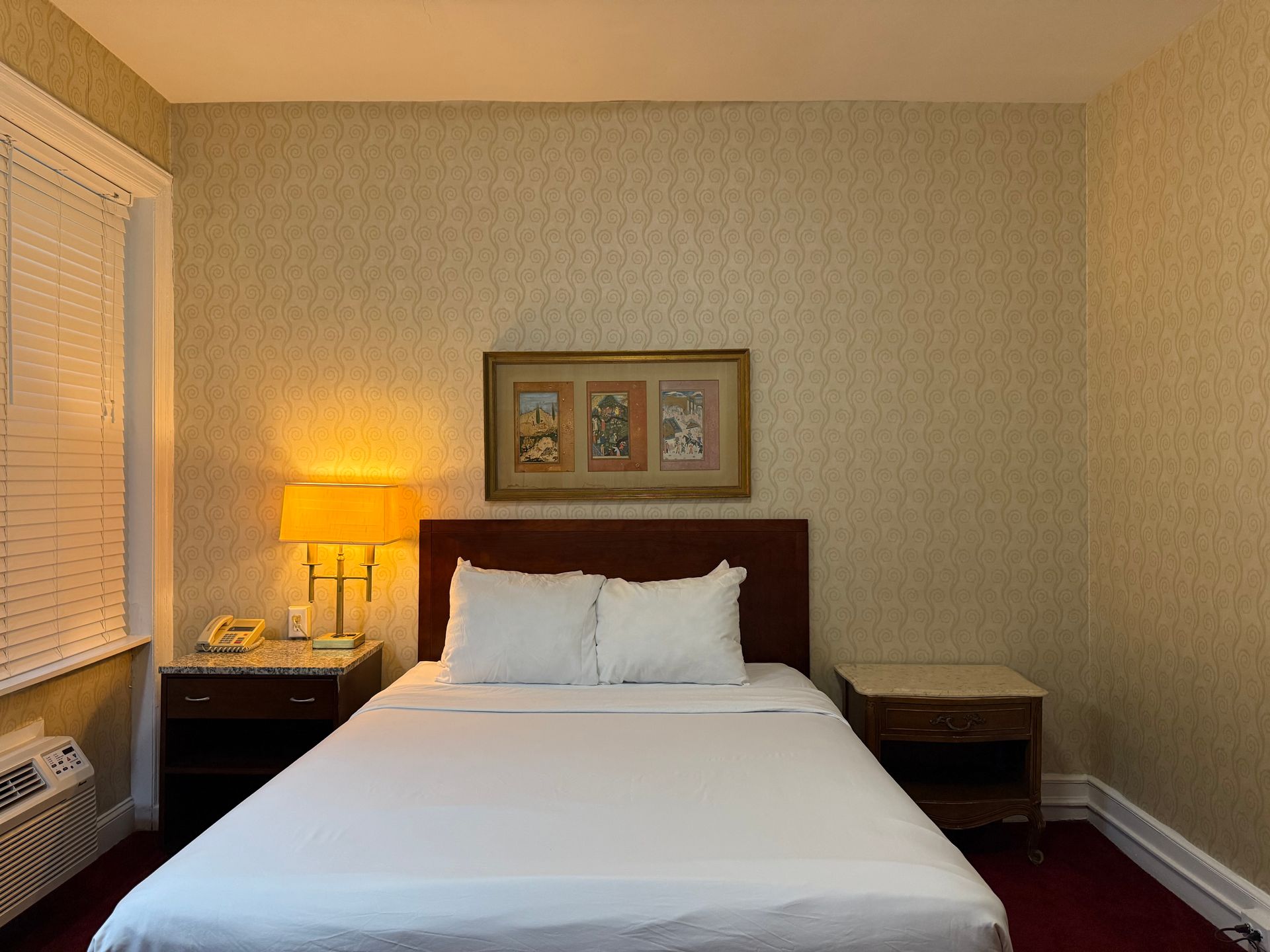 Bedroom with a made bed, nightstands, lamp, window with blinds, and a framed artwork.