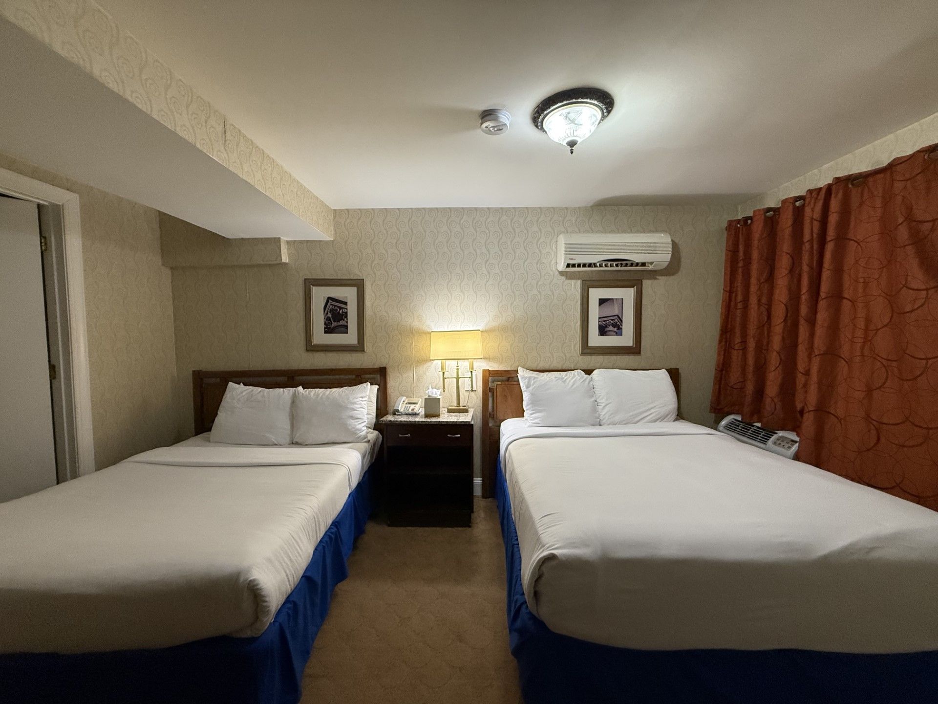 Two twin beds in a hotel room with bedside table, lamps, artwork, and orange curtains.