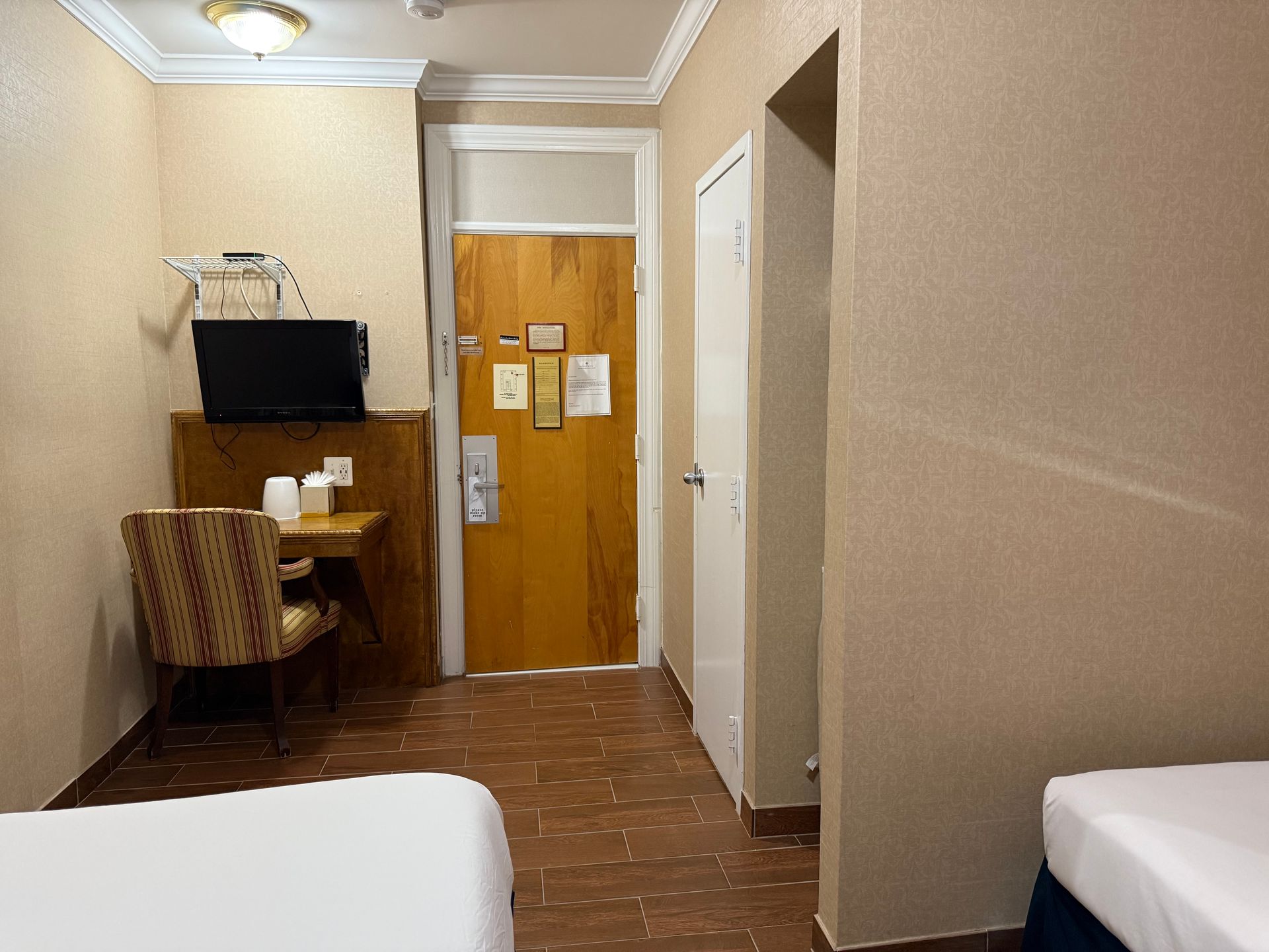 Hotel room with two beds, small desk, TV, and open doorway to hallway. Brown walls.