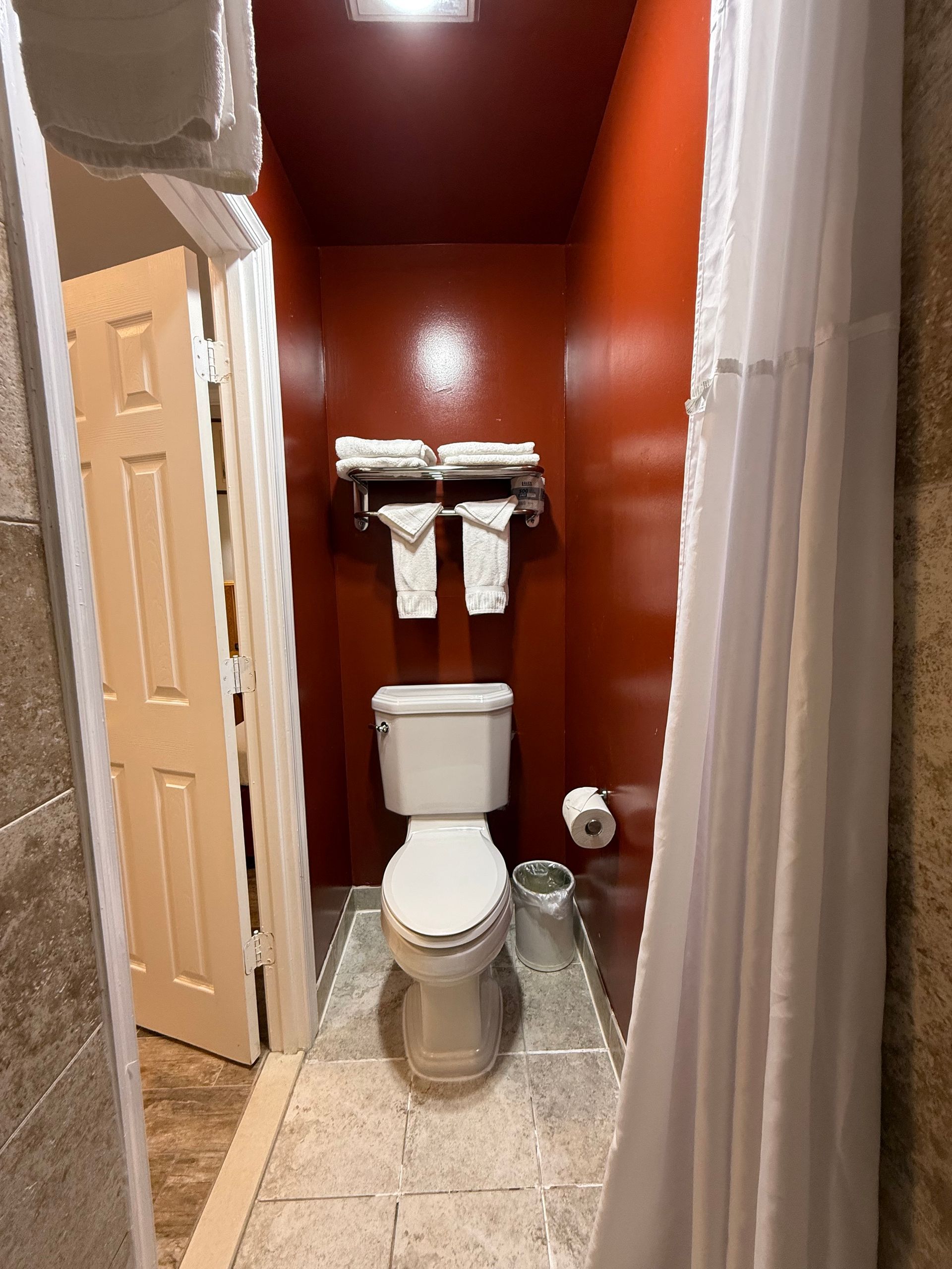 Small bathroom with red walls, toilet, and shower curtain. Towels and toilet paper are visible.