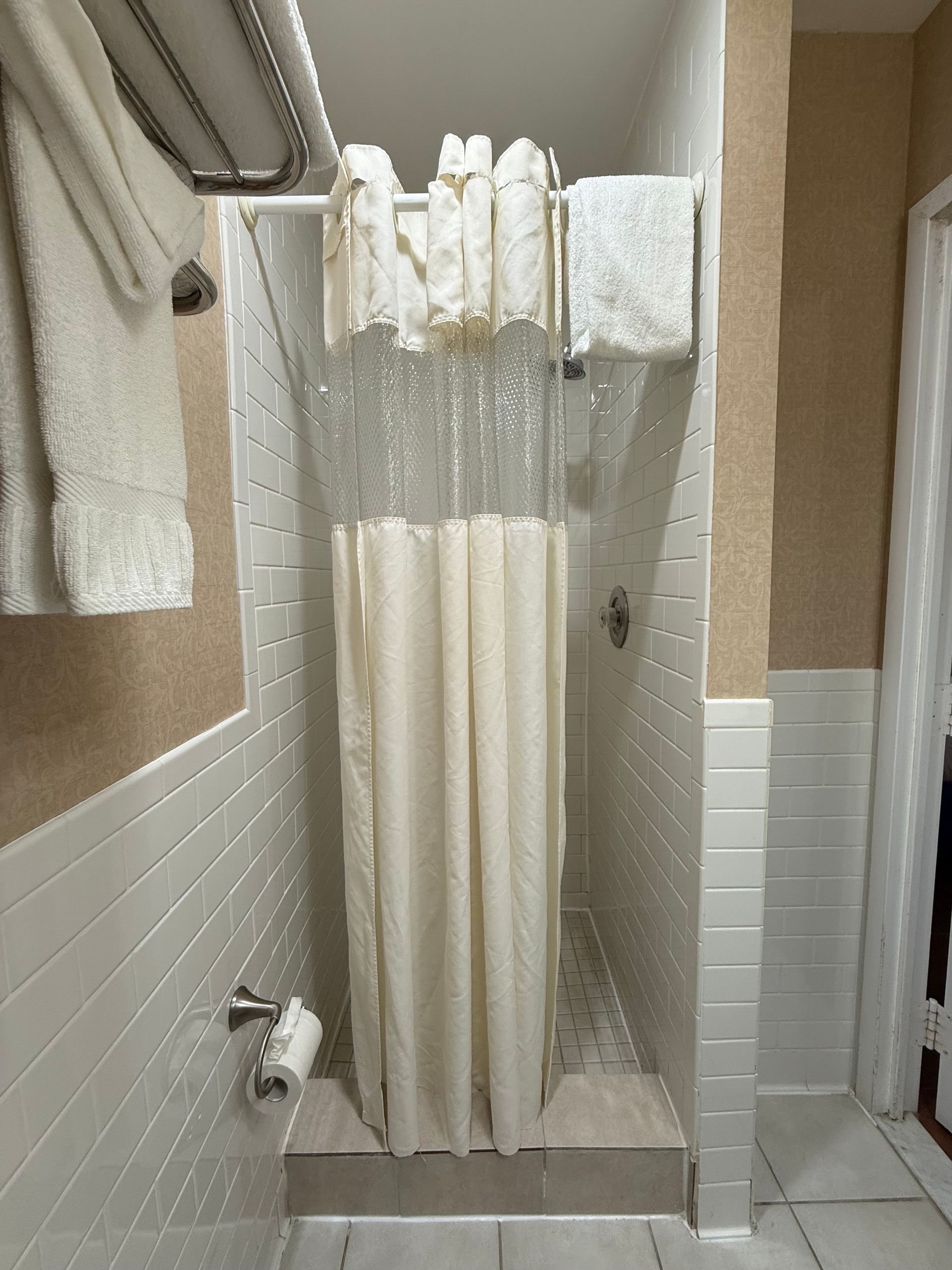 Narrow shower stall with a closed cream-colored shower curtain. White tiled walls, towels on a rack.