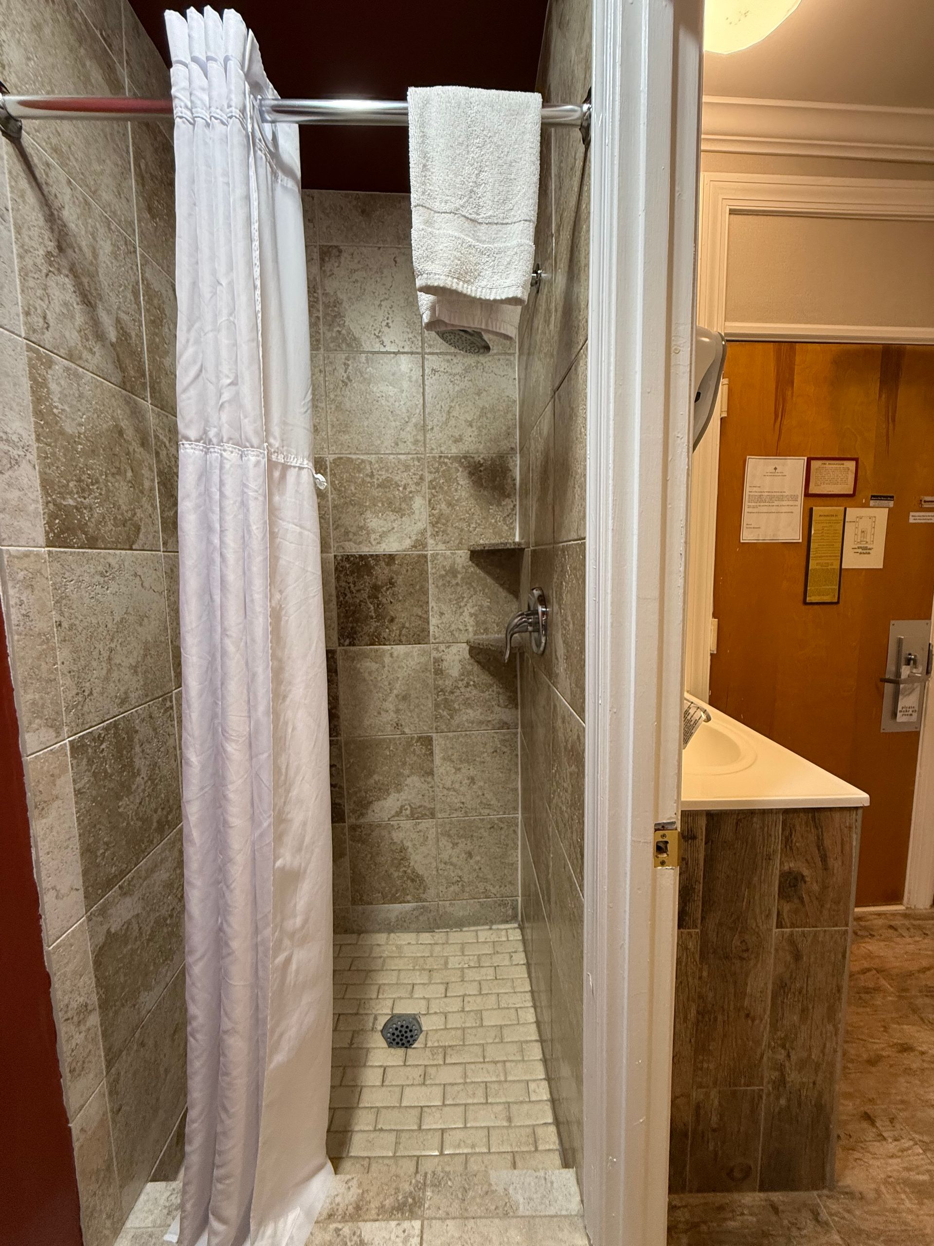 Shower stall with stone tile, white shower curtain, and a hanging towel.
