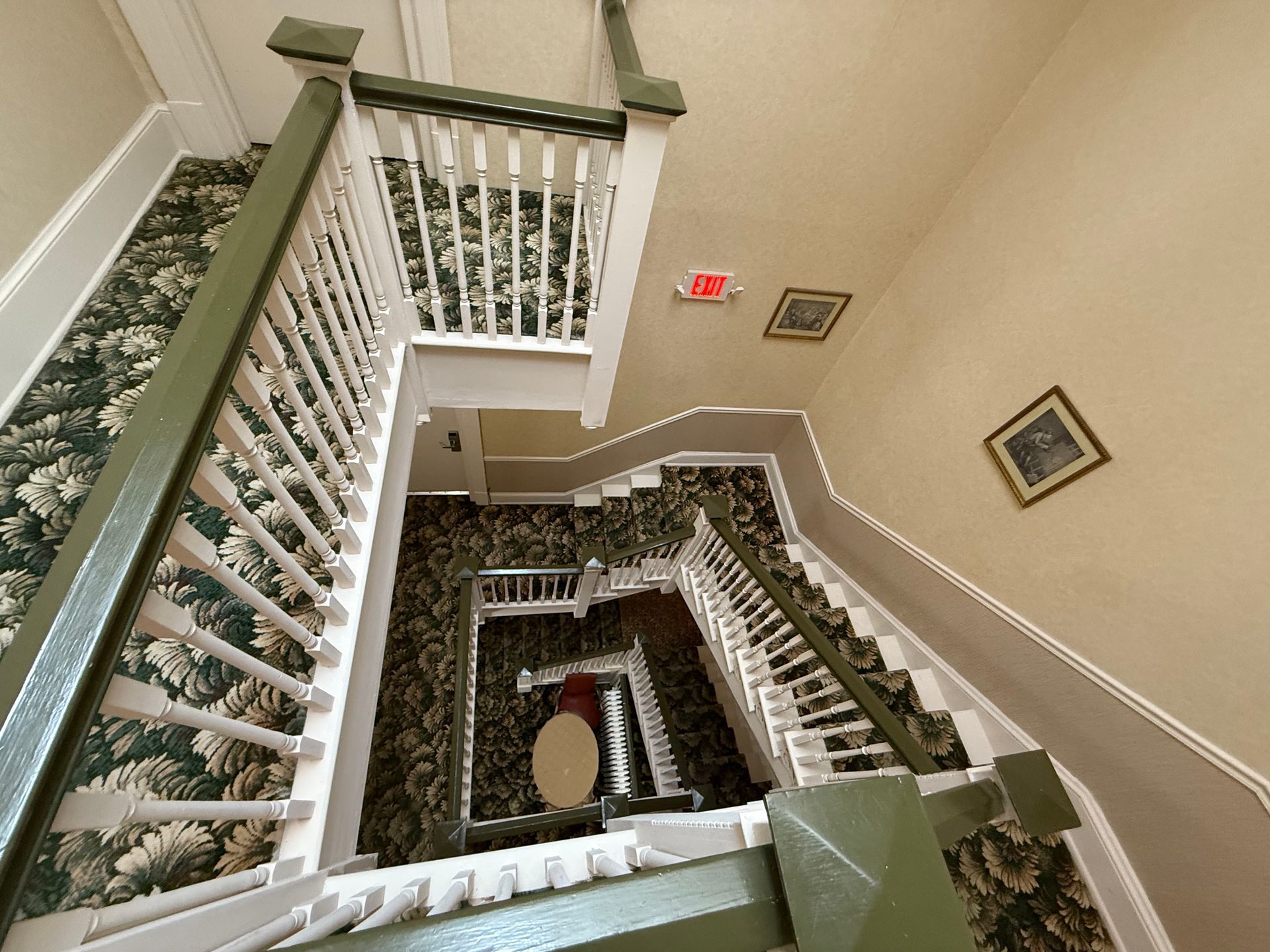 Looking down a winding staircase with white railings, green trim, floral wallpaper, and a red exit sign.