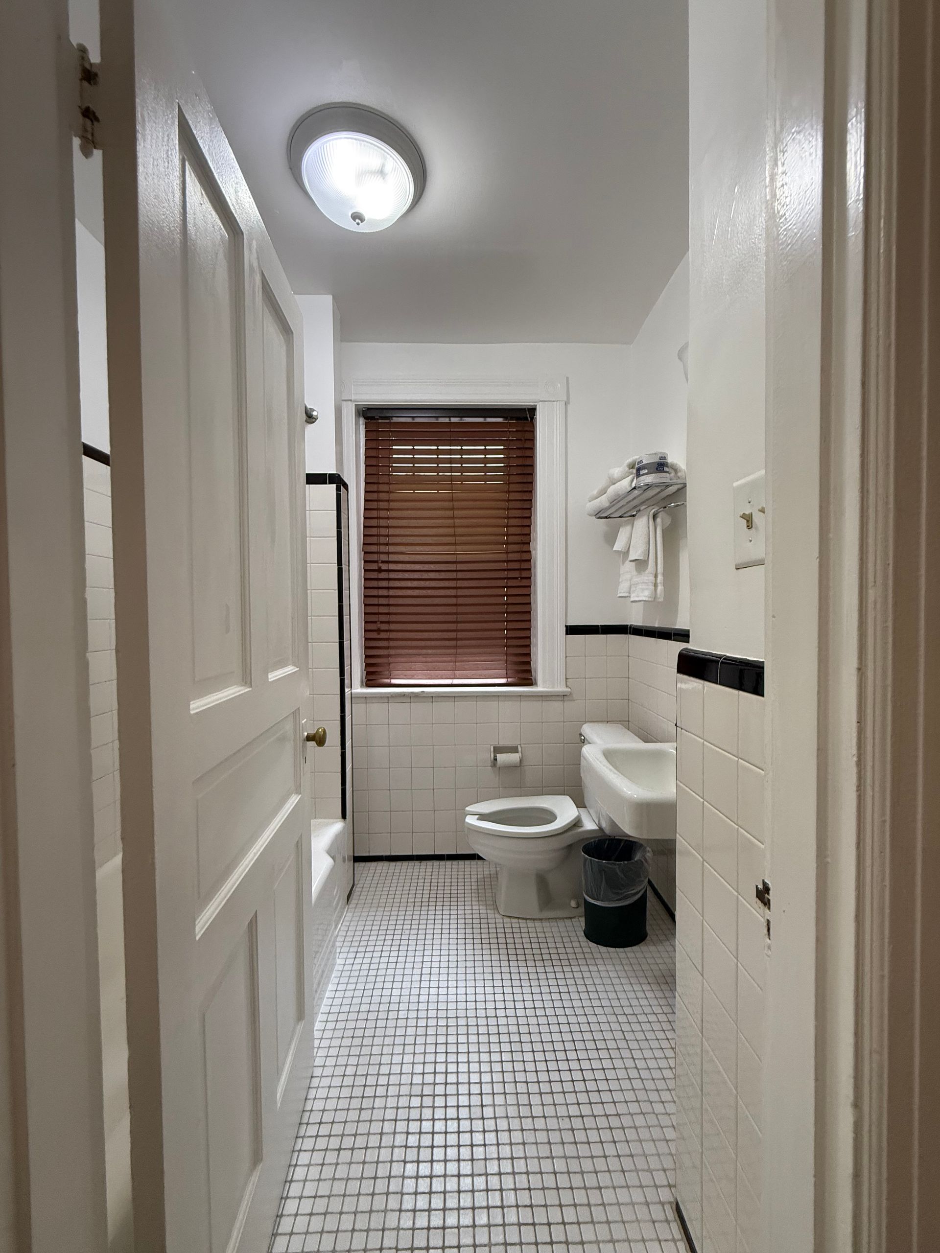 Bathroom with white tiles, black trim, patterned floor, toilet, sink, and window with blinds.