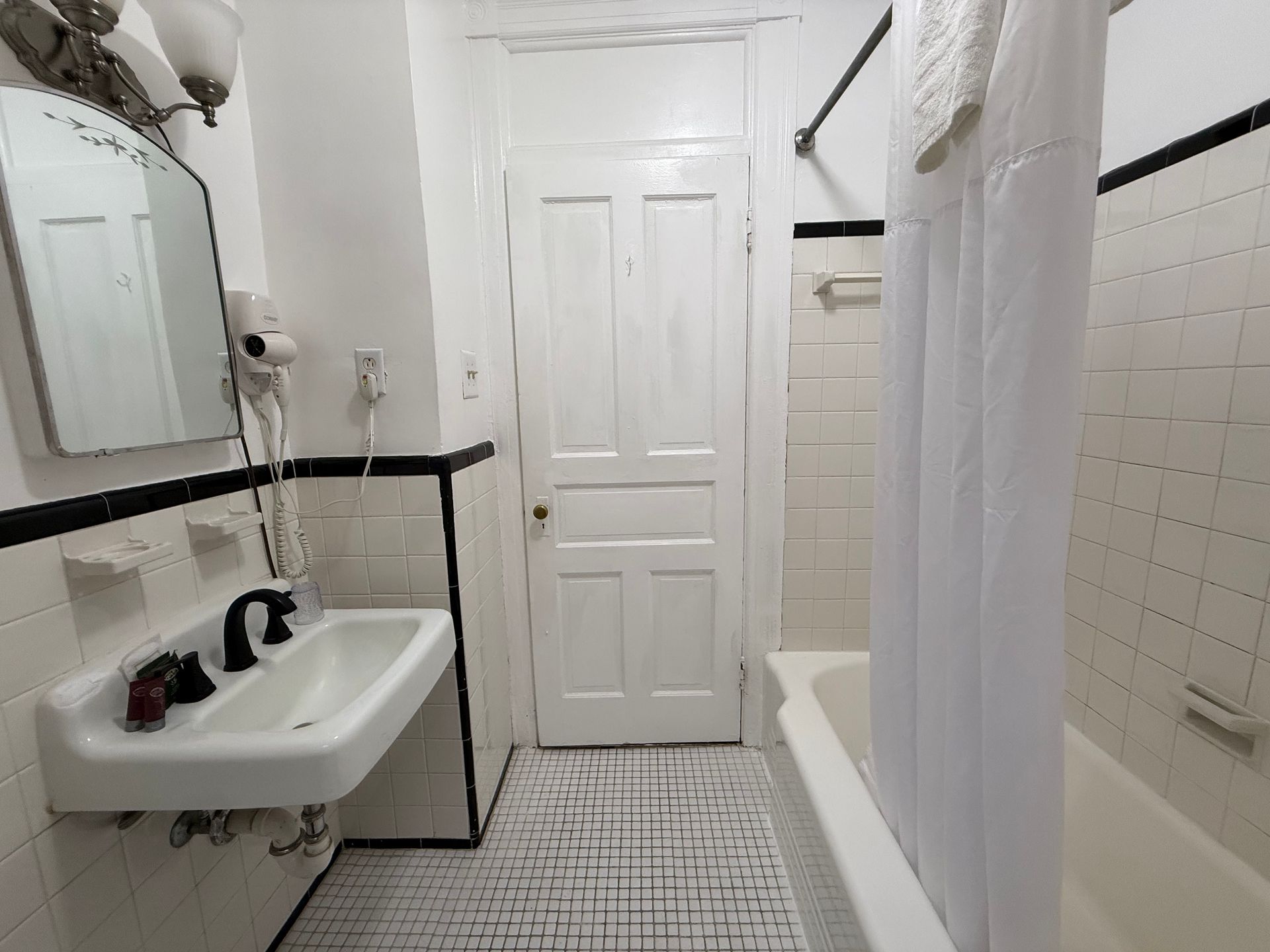 Small bathroom with white tile walls and a bathtub. Sink, mirror, and door visible.