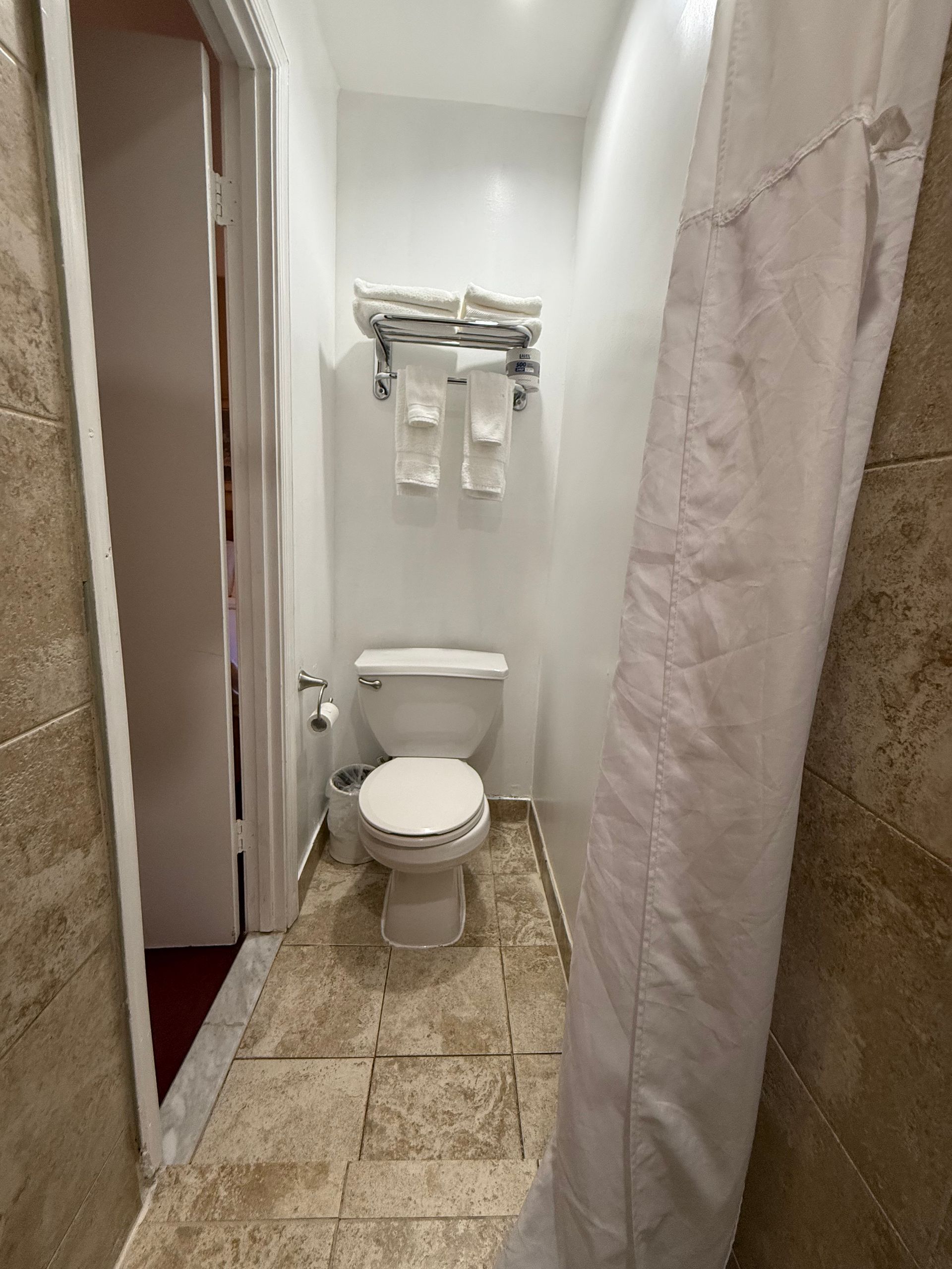 Small bathroom with toilet, towels, and shower curtain. Beige tile floor and white walls. Doorway to the left.