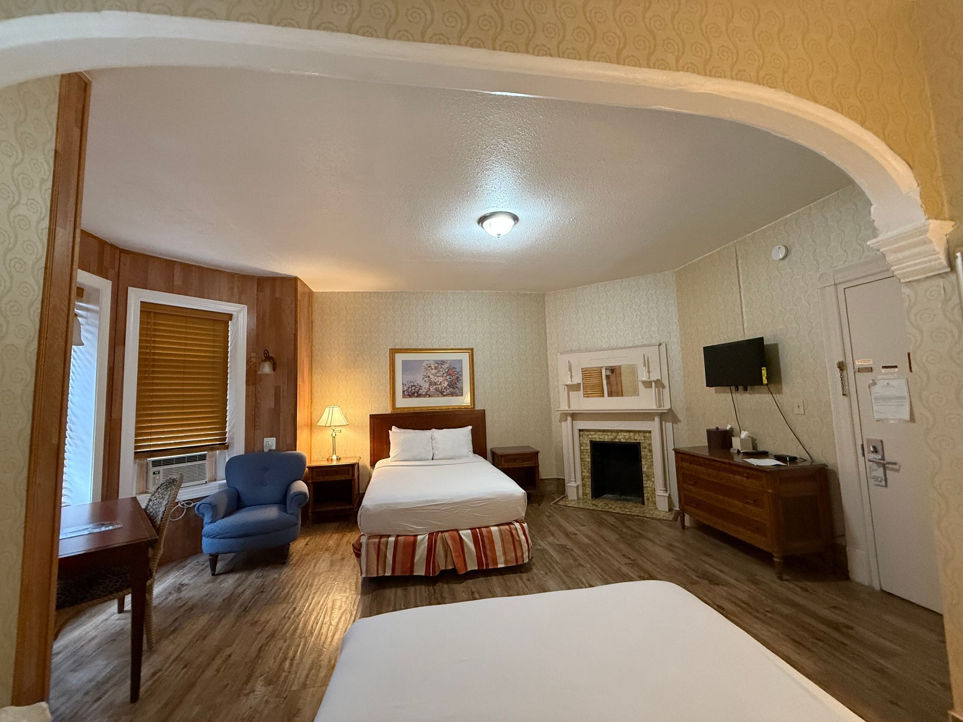 Hotel room with a bed, blue chair, fireplace, tv, and desk. The walls are light.